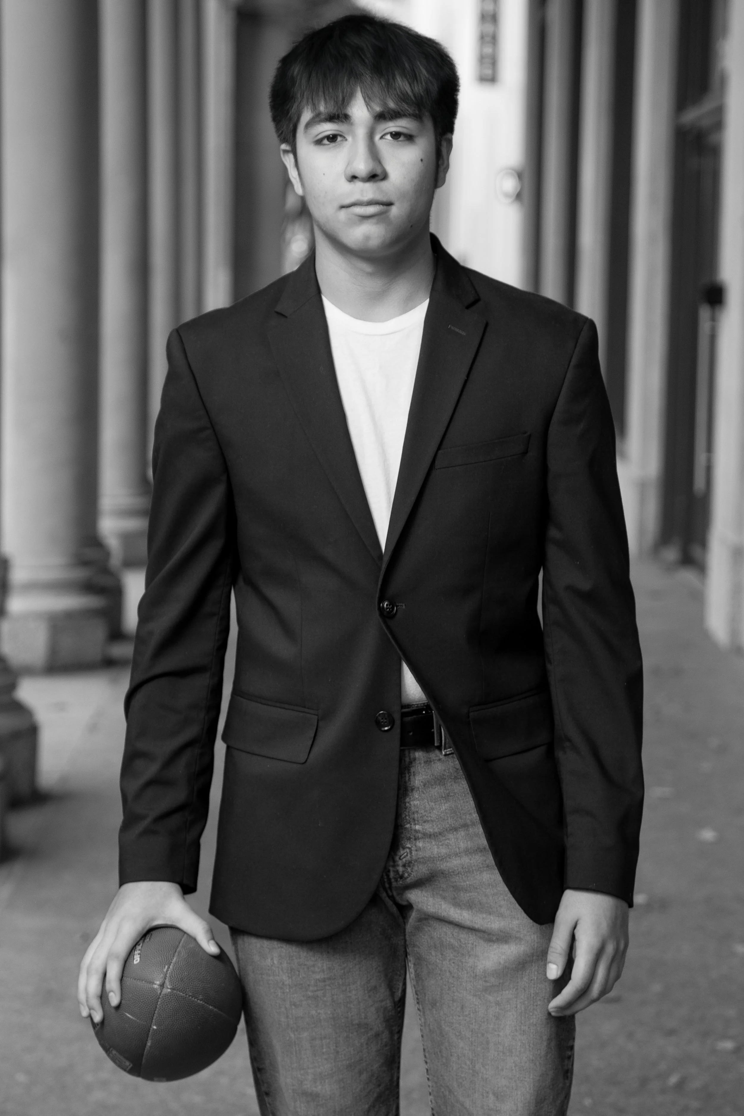 Black and white high school senior portrait featuring a male senior in a suit holding a football in a classic urban setting. Timeless, editorial-style senior photography by Dominic Alvarez Photography serving Houston, Katy, and surrounding areas acro
