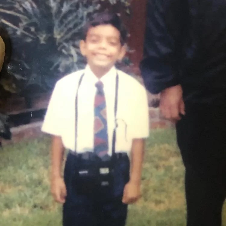 A young boy smiling outdoors, wearing a white shirt, a colorful tie, and holding a camera with a strap around his neck. An adult's arm is partially visible beside him.