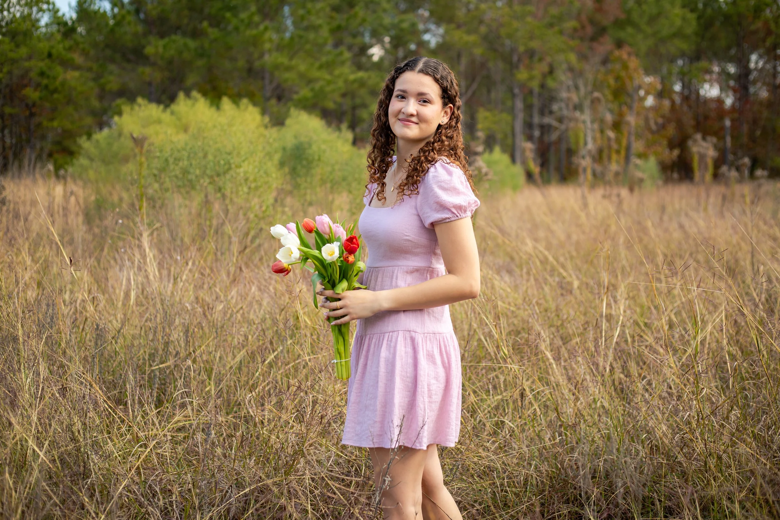 Natural light outdoor high school senior portrait of a teenage girl in a pink dress holding tulips in a field with trees in the background. Soft, bright, and timeless senior photography in the Houston, Texas area.