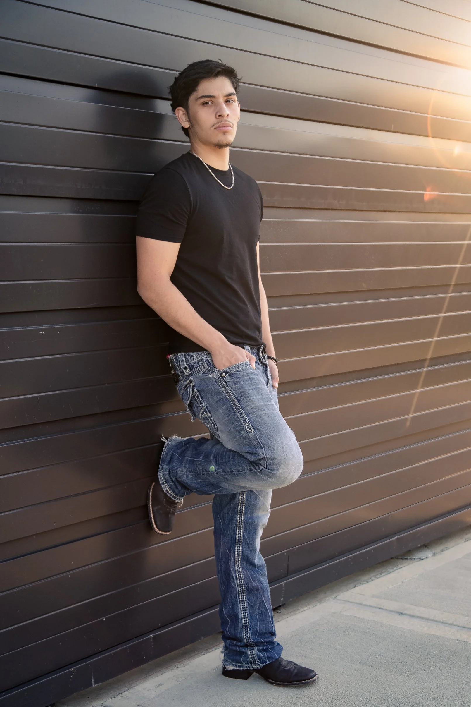 A Houston high school senior young man wearing a black t-shirt, jeans, and black boots leaning against a brown wall with his hands in his pockets. He is standing outdoors during sunset with sunlight flare visible.