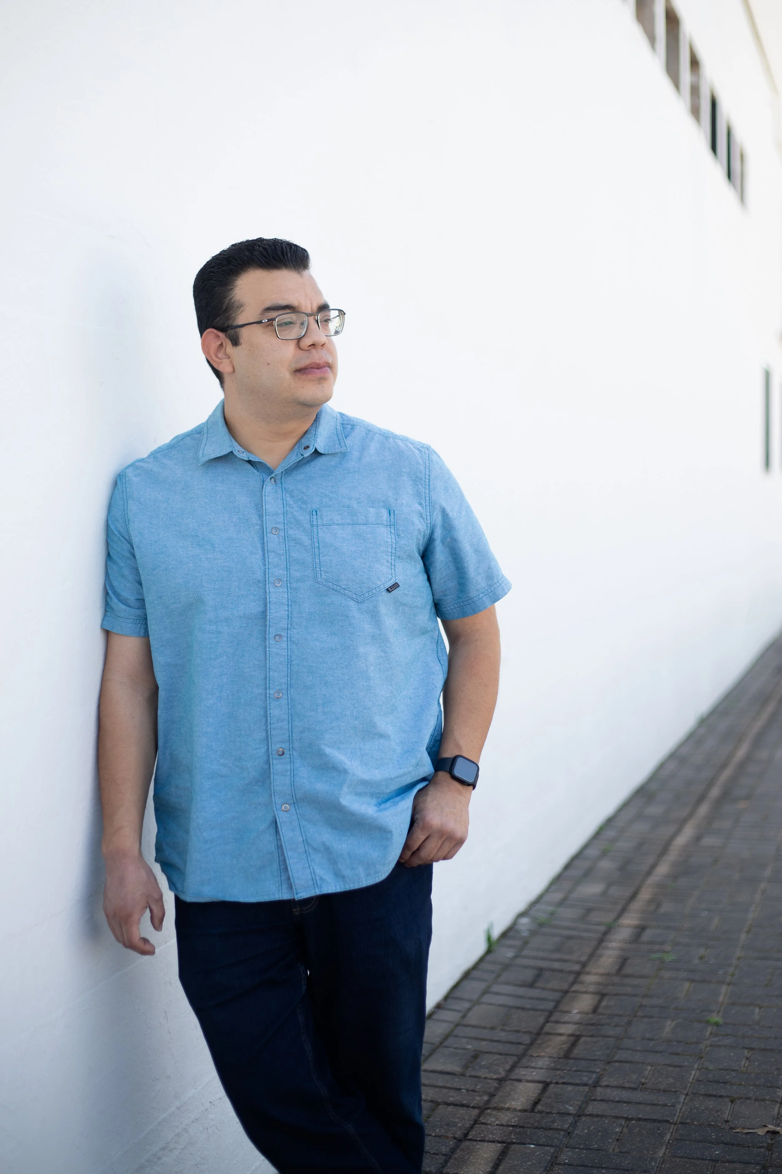 A man in glasses, a blue short-sleeve button-down shirt, and dark jeans stands outdoors against a white wall. He has his eyes closed and his right hand in his pocket.