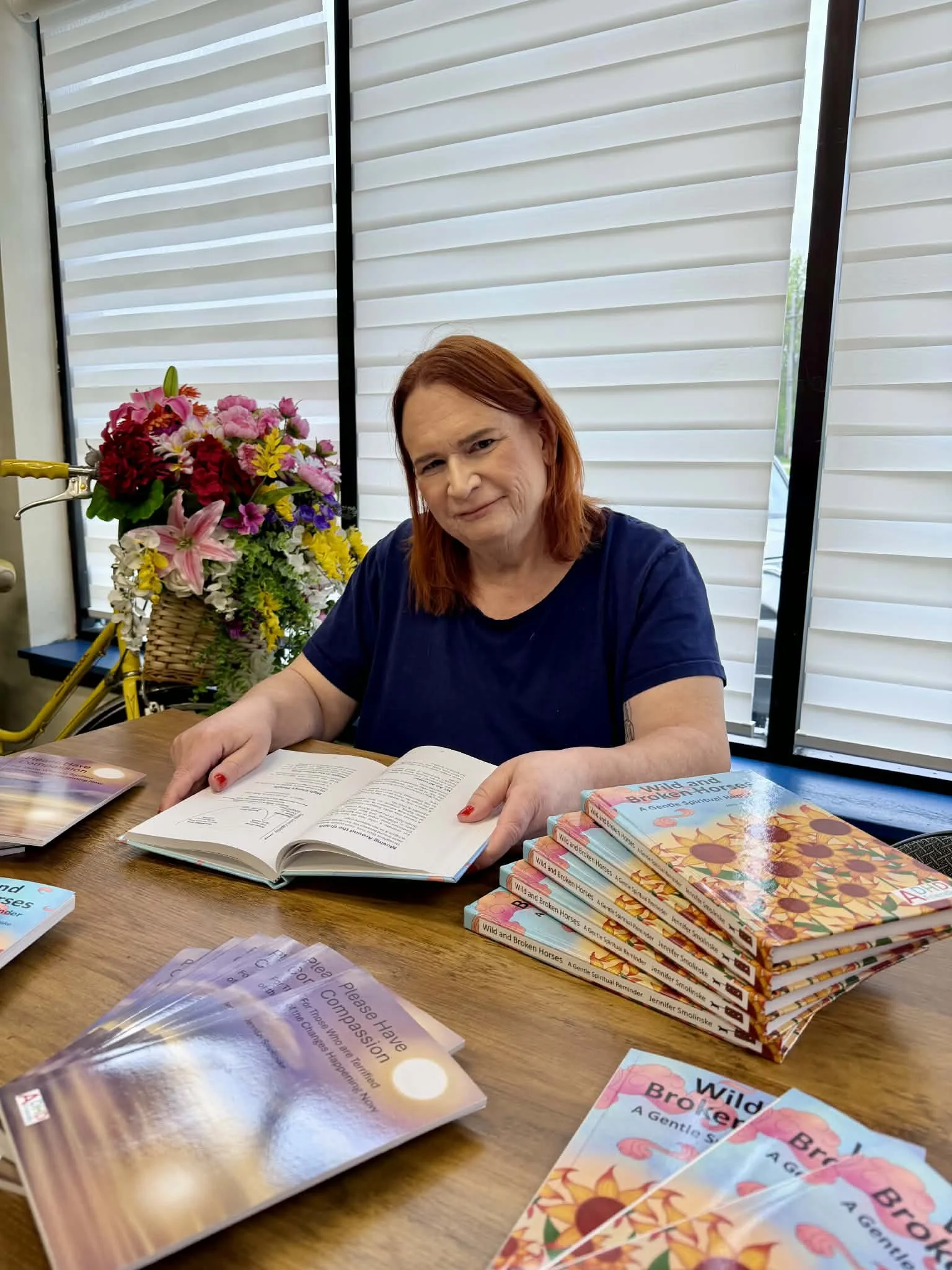A woman with red hair sitting at a wooden table with books, pamphlets, and a bouquet of colorful flowers. The background shows modern window blinds.