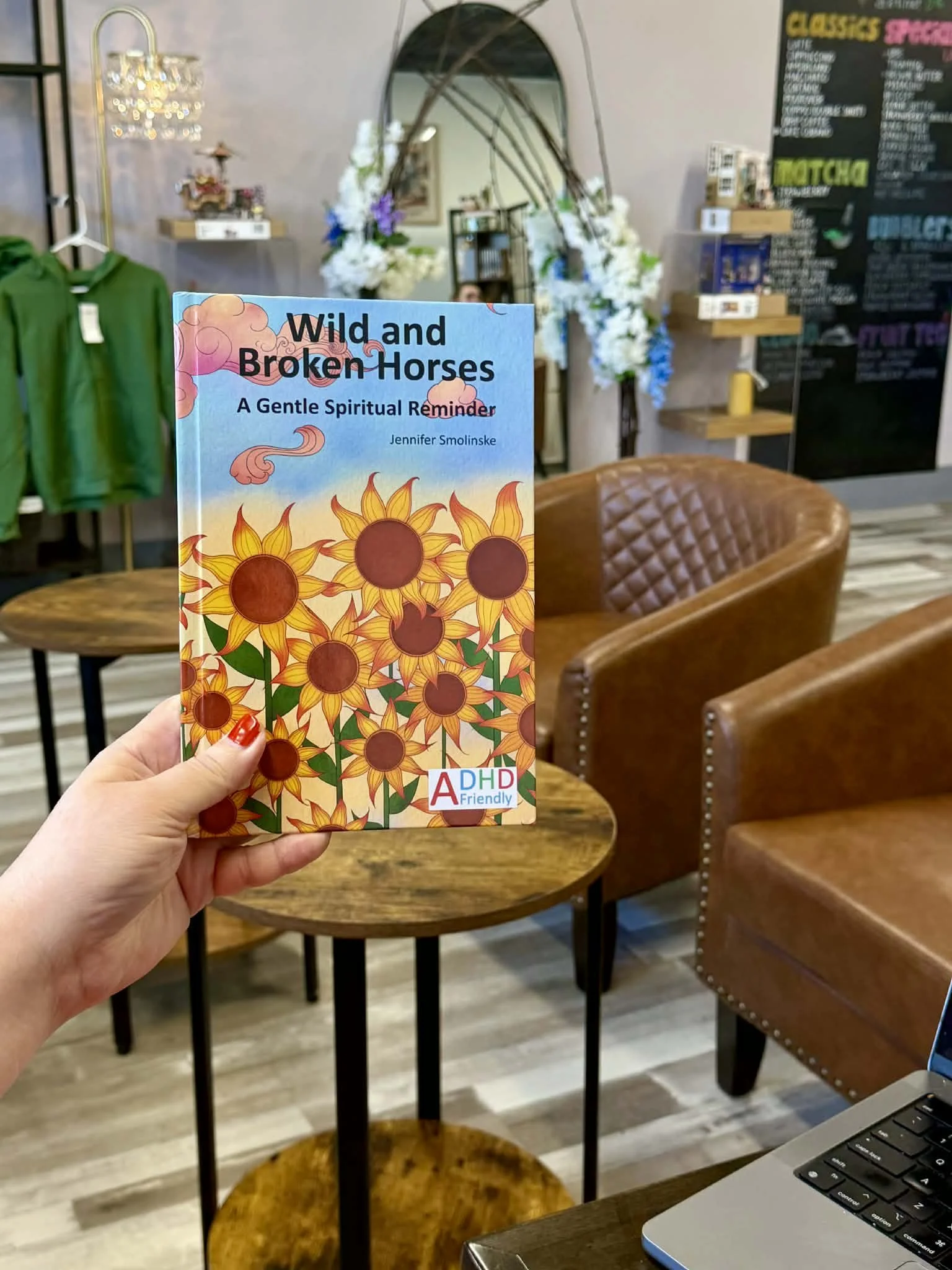A person holding a book titled "Wild and Broken Horses: A Gentle Spiritual Reminder" with sunflower illustration on the cover, inside a cafe with wooden tables, brown chairs, a blackboard menu, and floral decorations in the background.