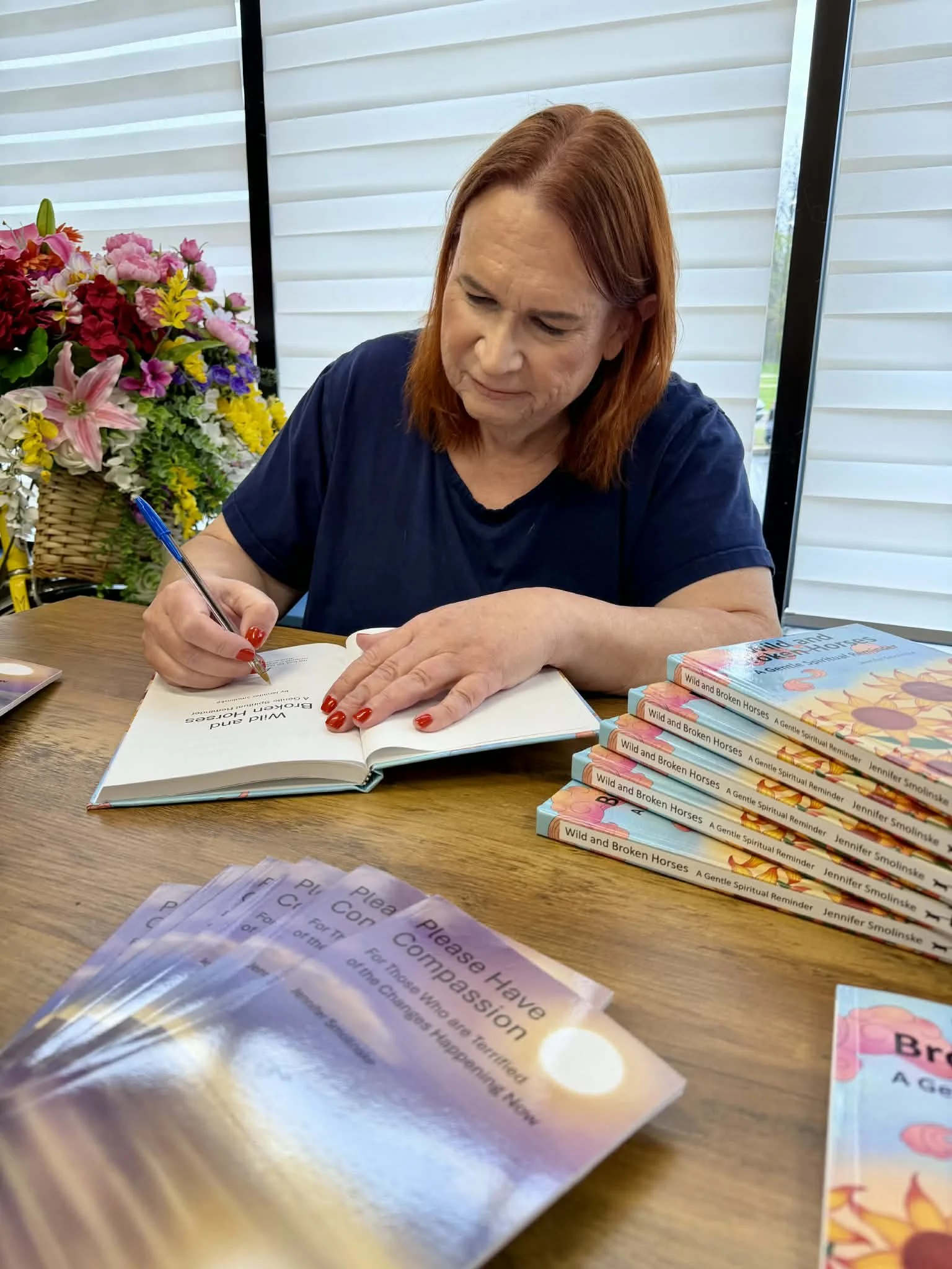 A woman with red hair is signing a book titled "Wild and Broken Horses" by Jennifer Smolinske, with several copies stacked on the table beside her. There is a large bouquet of flowers in a basket in the background and some pamphlets in the foreground.