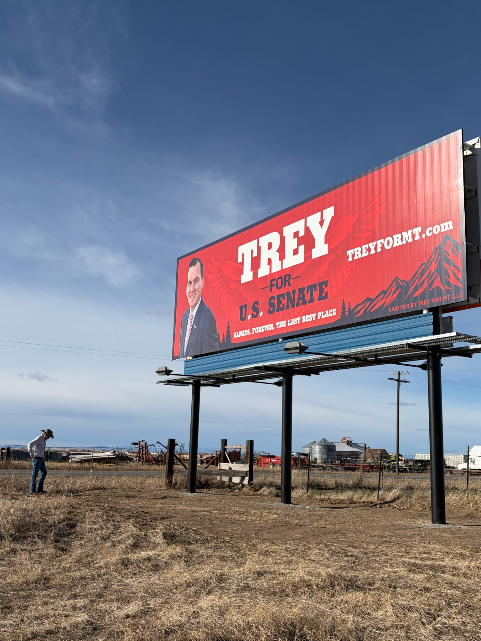 Billboard advertising Trey for U.S. Senate with a man in a cowboy hat standing on dry grass patch, farm equipment, buildings, and a blue sky in the background.