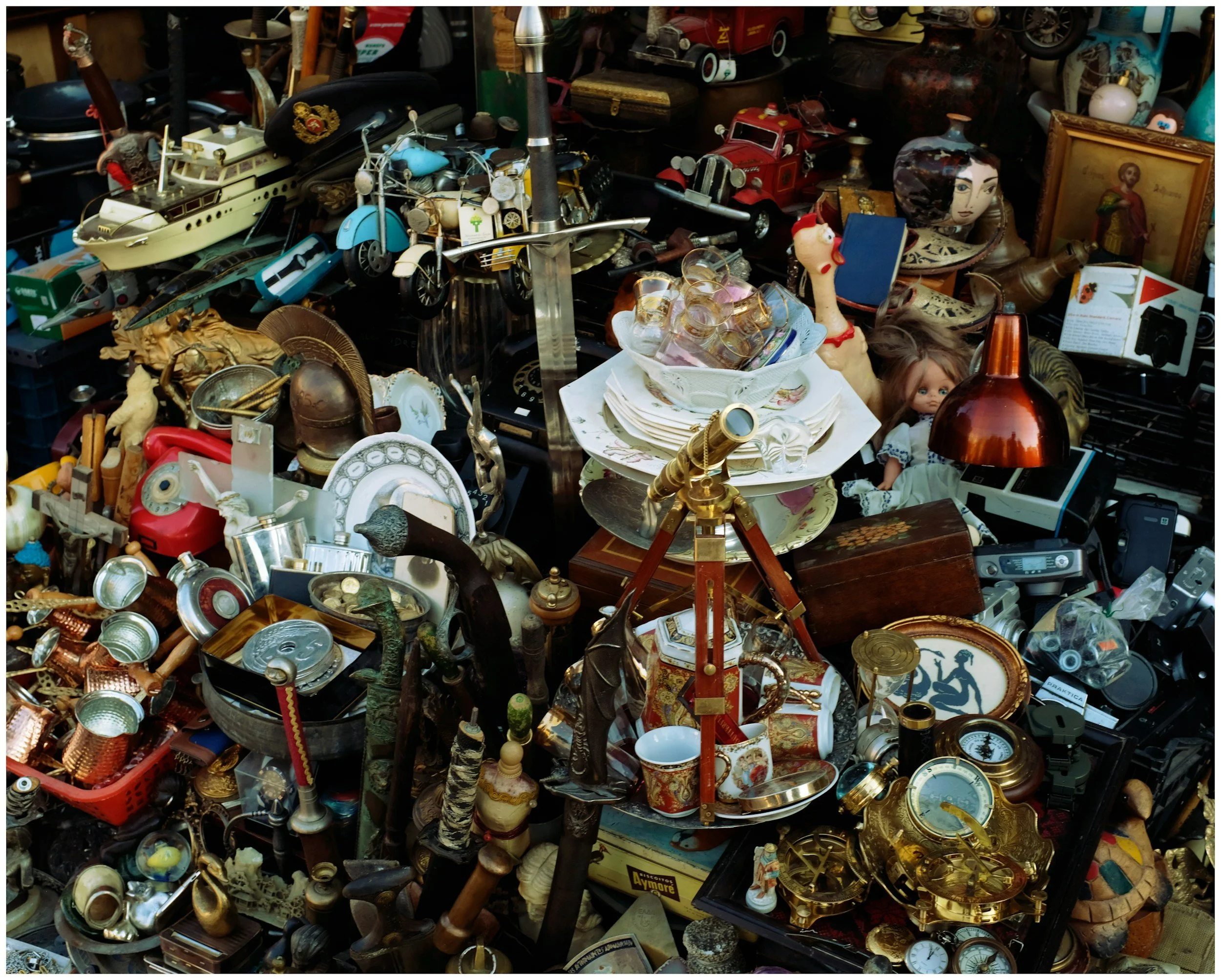 A collection of vintage and antique items including small toy cars, doll, old lamps, brass and metal objects, cameras, and various trinkets displayed on a table or stall.