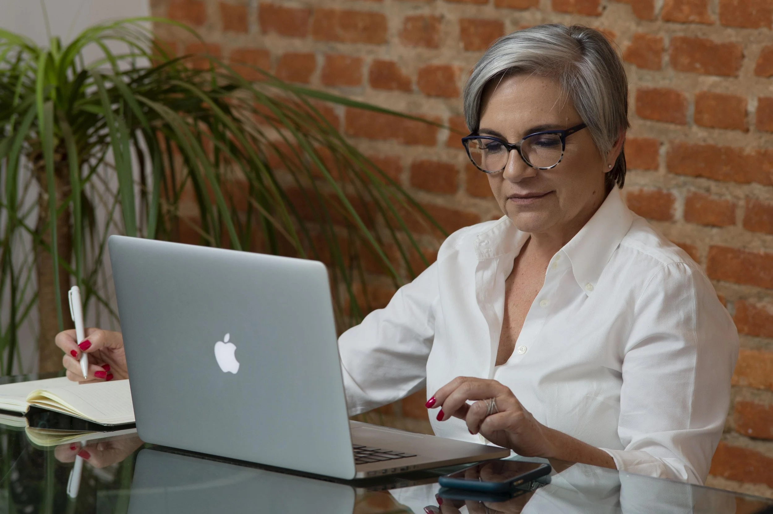 Mulher com óculos usando um laptop prateado em um ambiente com parede de tijolos, segurando uma caneta na mão direita e uma folha de papel amarela na esquerda, com um celular em cima da mesa.