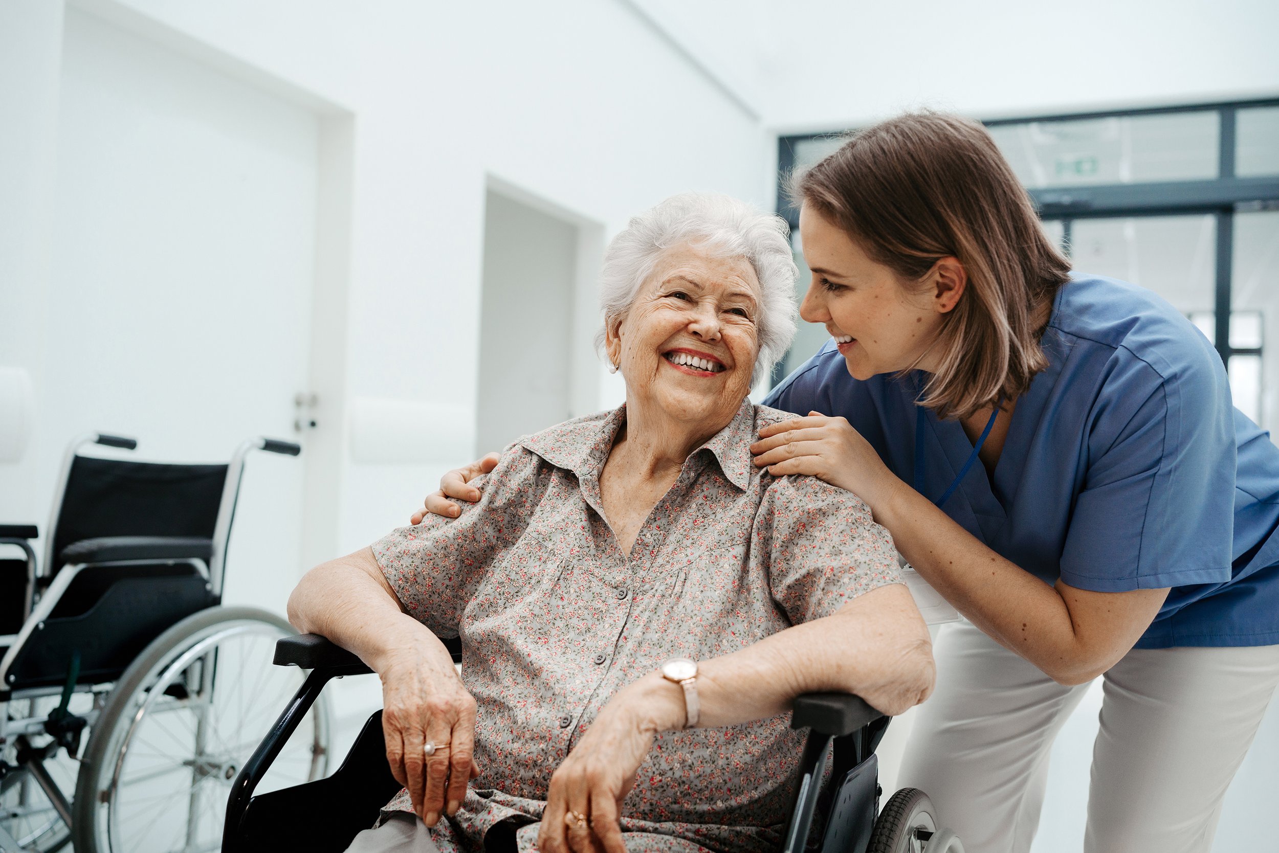 Nurse or caregiver smiling and talking with elderly woman in wheelchair, both showing a caring and joyful interaction in a bright, modern healthcare facility.