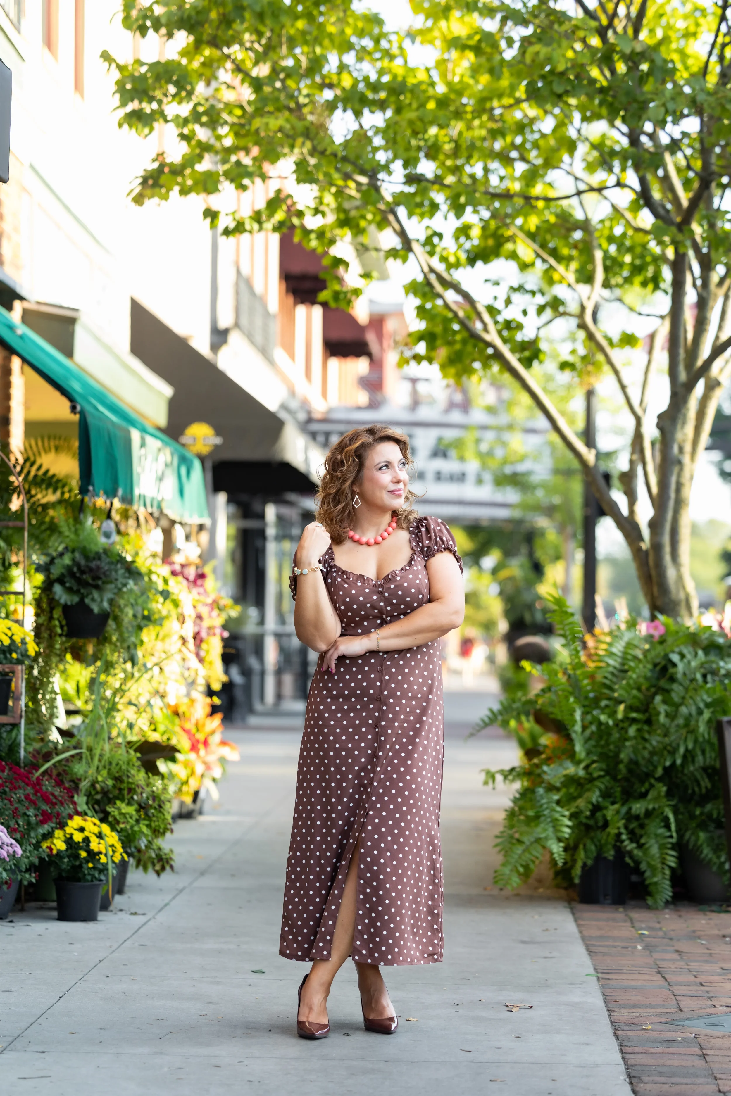A woman stands on a city sidewalk lined with potted plants and flowers, wearing a brown polka dot dress, with trees and storefronts in the background.