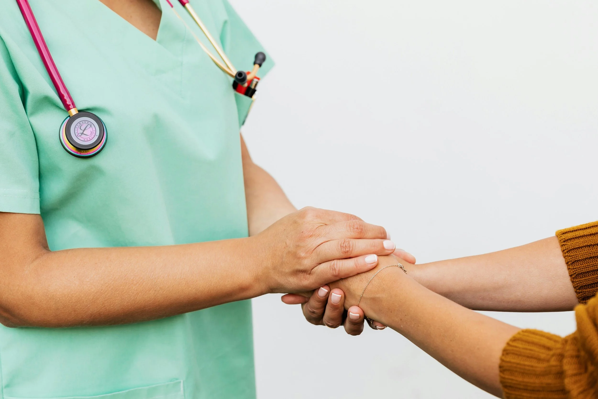 Healthcare professional in green scrubs shaking hands with a patient, who is wearing a mustard-colored sweater, against a plain white background.