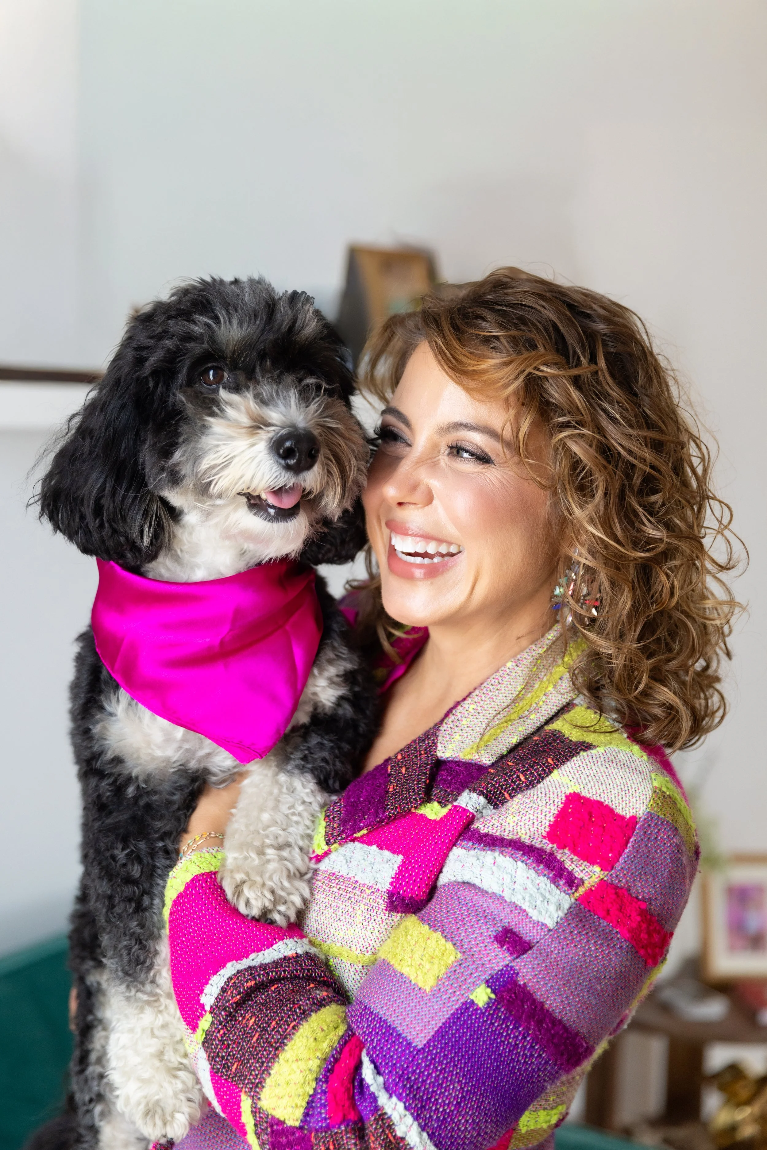 A woman with curly hair smiling and holding a black and white dog wearing a bright pink bandana.