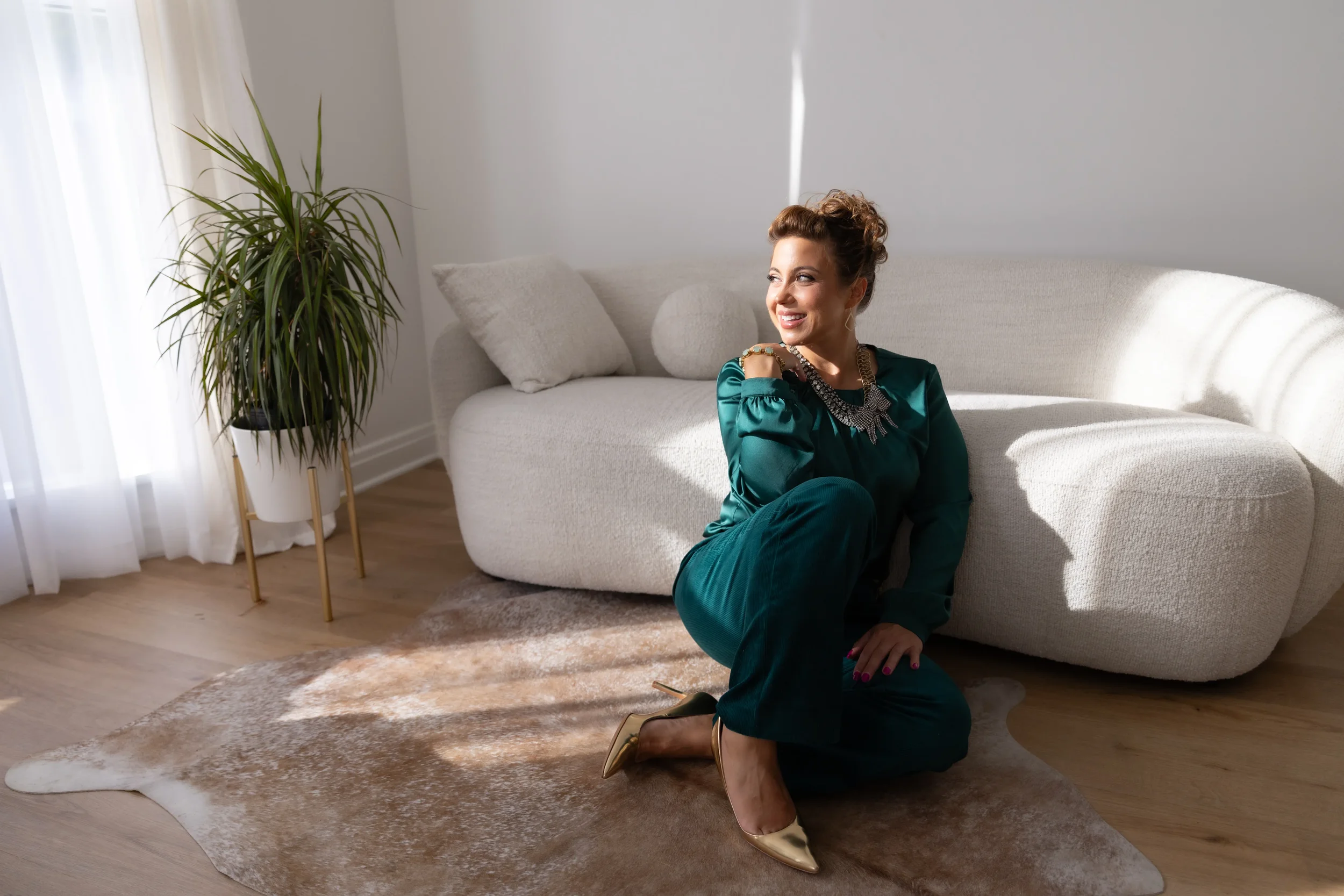 A woman in teal outfit with gold shoes and statement necklace, sitting on her knees on a cowhide rug in a bright, modern living room, smiling and looking to her left.