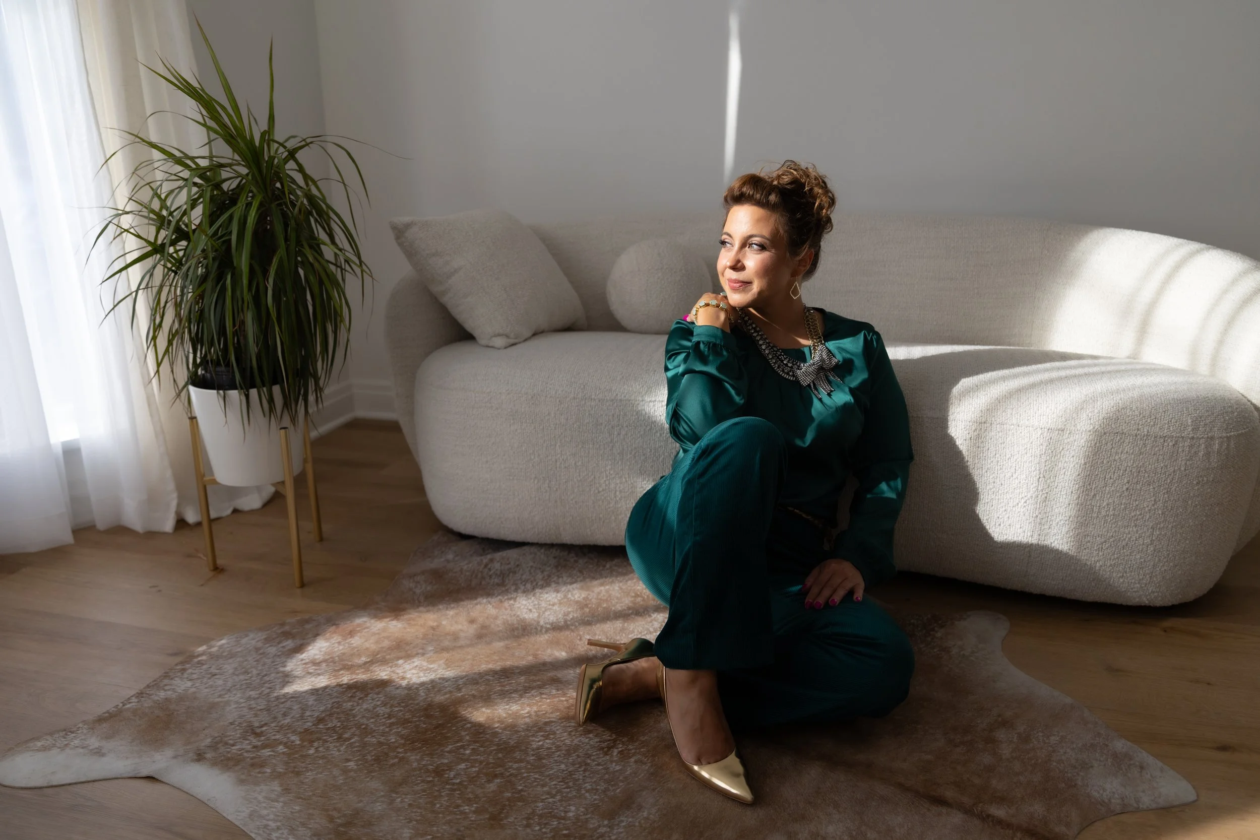 A woman in dark green attire sitting on a cowhide rug in a living room, with a beige sofa and a tall potted plant nearby, illuminated by sunlight.