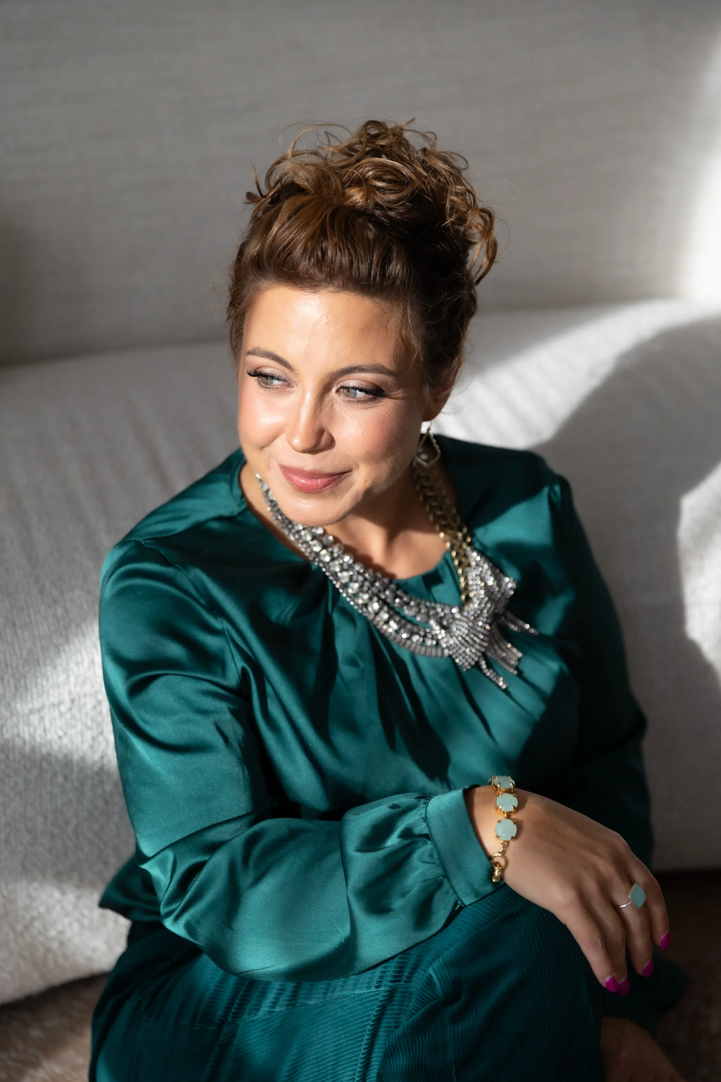 Woman with brown hair styled in an updo, wearing a teal satin blouse and adorned with a large silver statement necklace, sitting on a light-colored sofa in natural light.