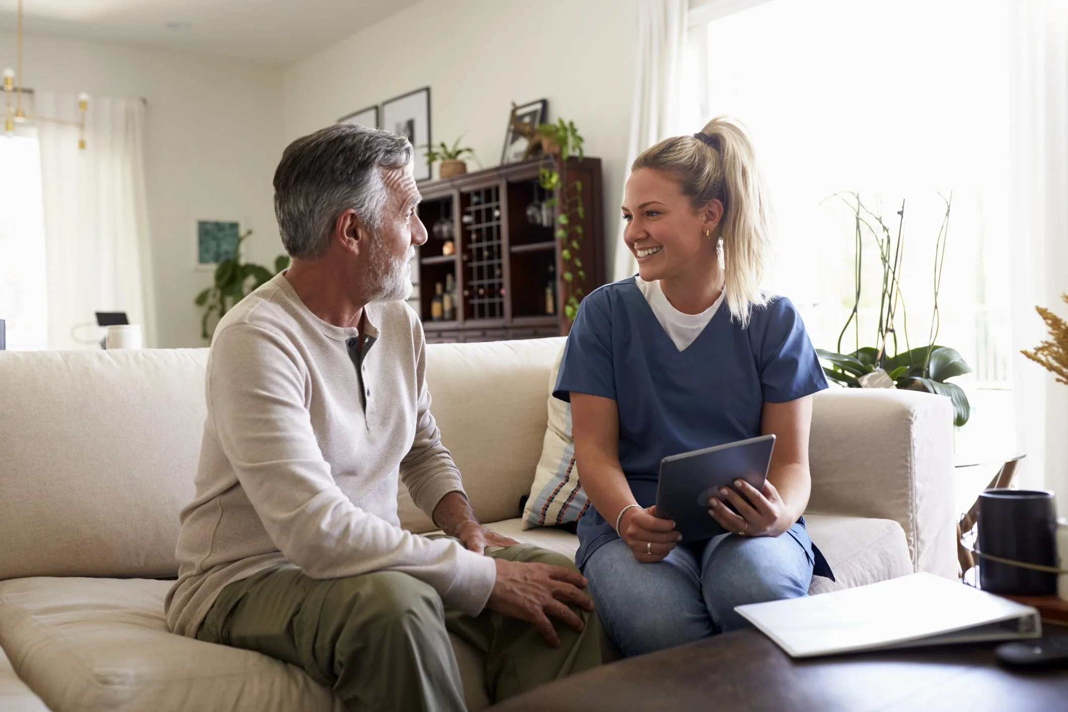 A young woman in medical scrubs sitting on a beige couch, smiling and holding a tablet, talking to an older man with gray hair and a beard, also sitting on the couch, in a well-lit living room.