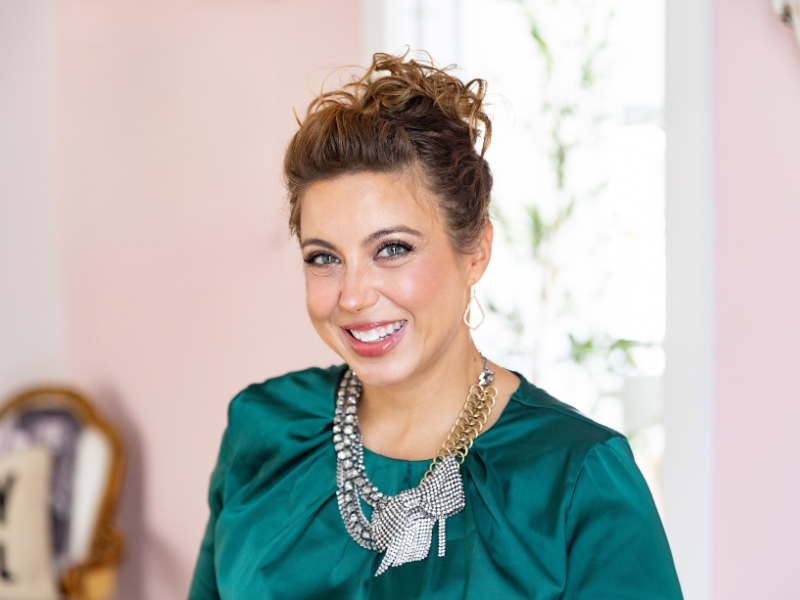 A smiling woman with brown hair styled in an updo, wearing a teal blouse and a large statement necklace, standing in a room with pink walls and a window behind her.