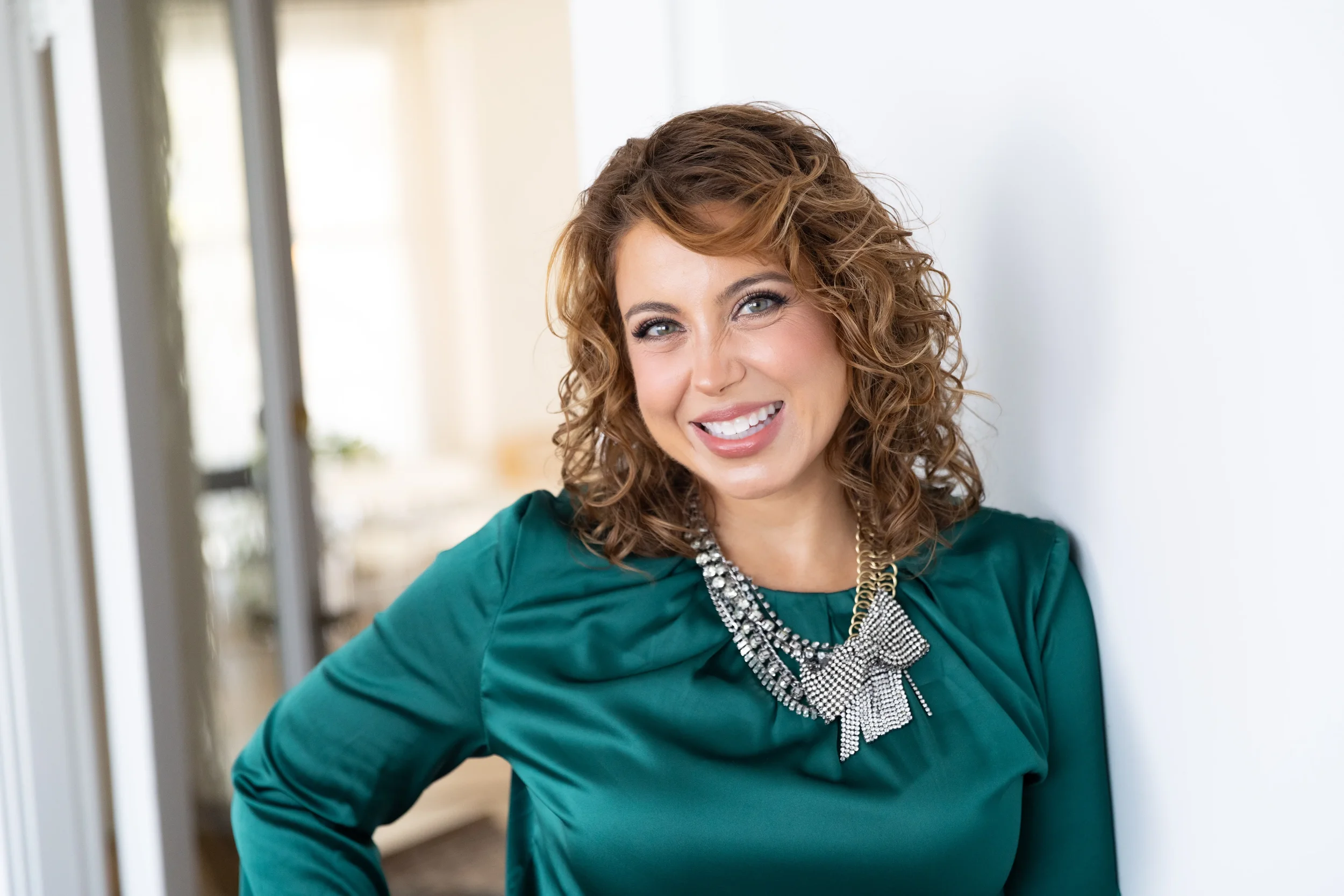 A woman with curly brown hair smiling and leaning against a white wall, wearing a teal top and a statement necklace.