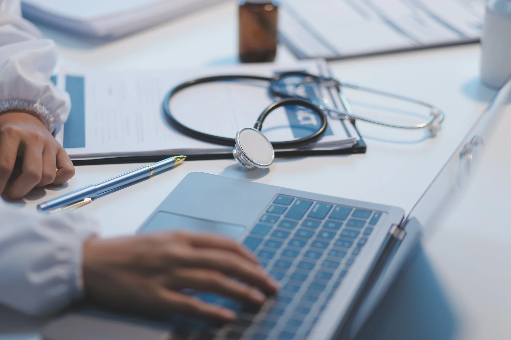 A medical workspace on a white desk with a stethoscope, a pen, a laptop, and medication bottles in the background.