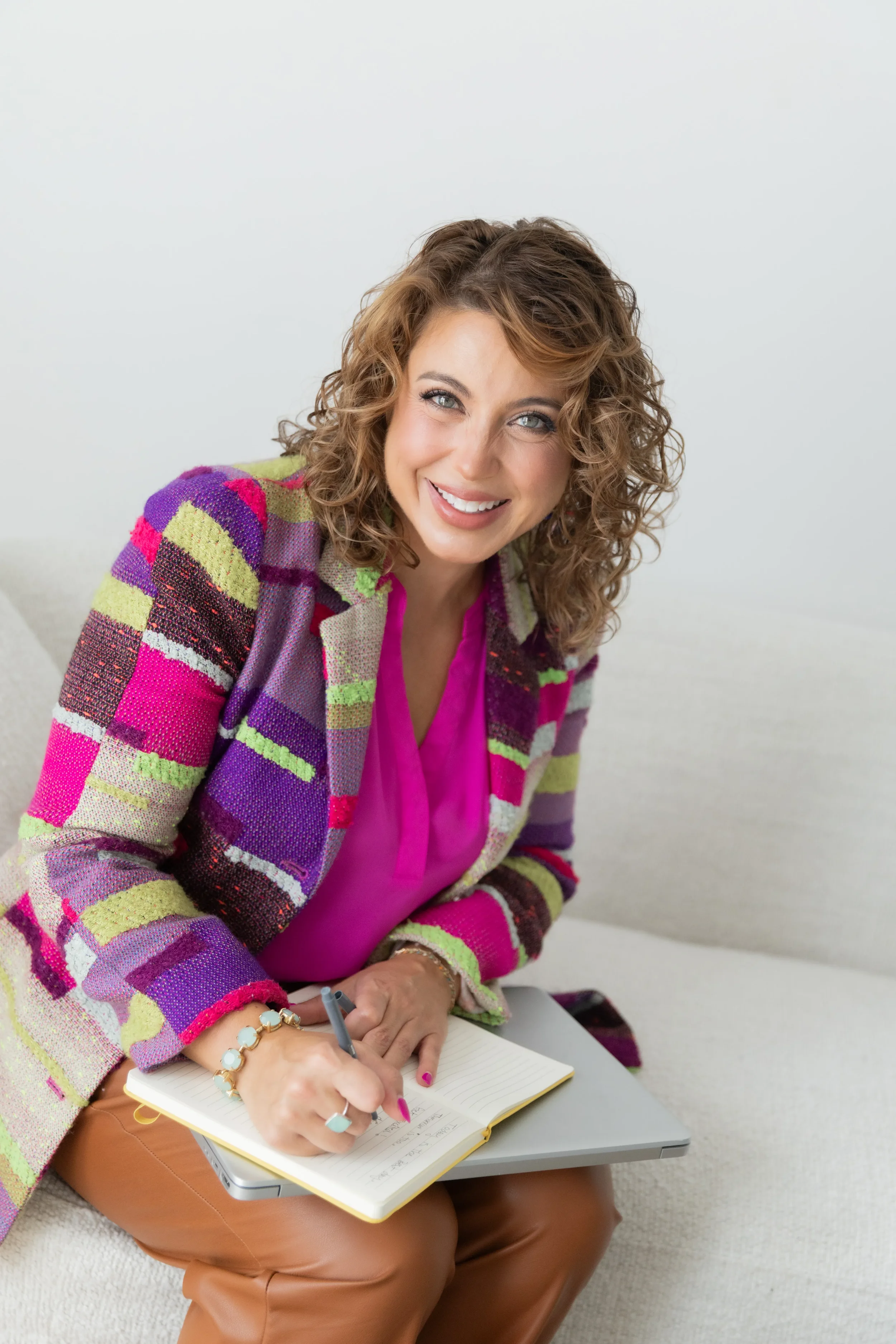 A woman with curly hair, wearing a colorful plaid blazer and pink blouse, sitting on a beige sofa, smiling, and writing in a yellow notebook placed on her lap with a laptop next to her.
