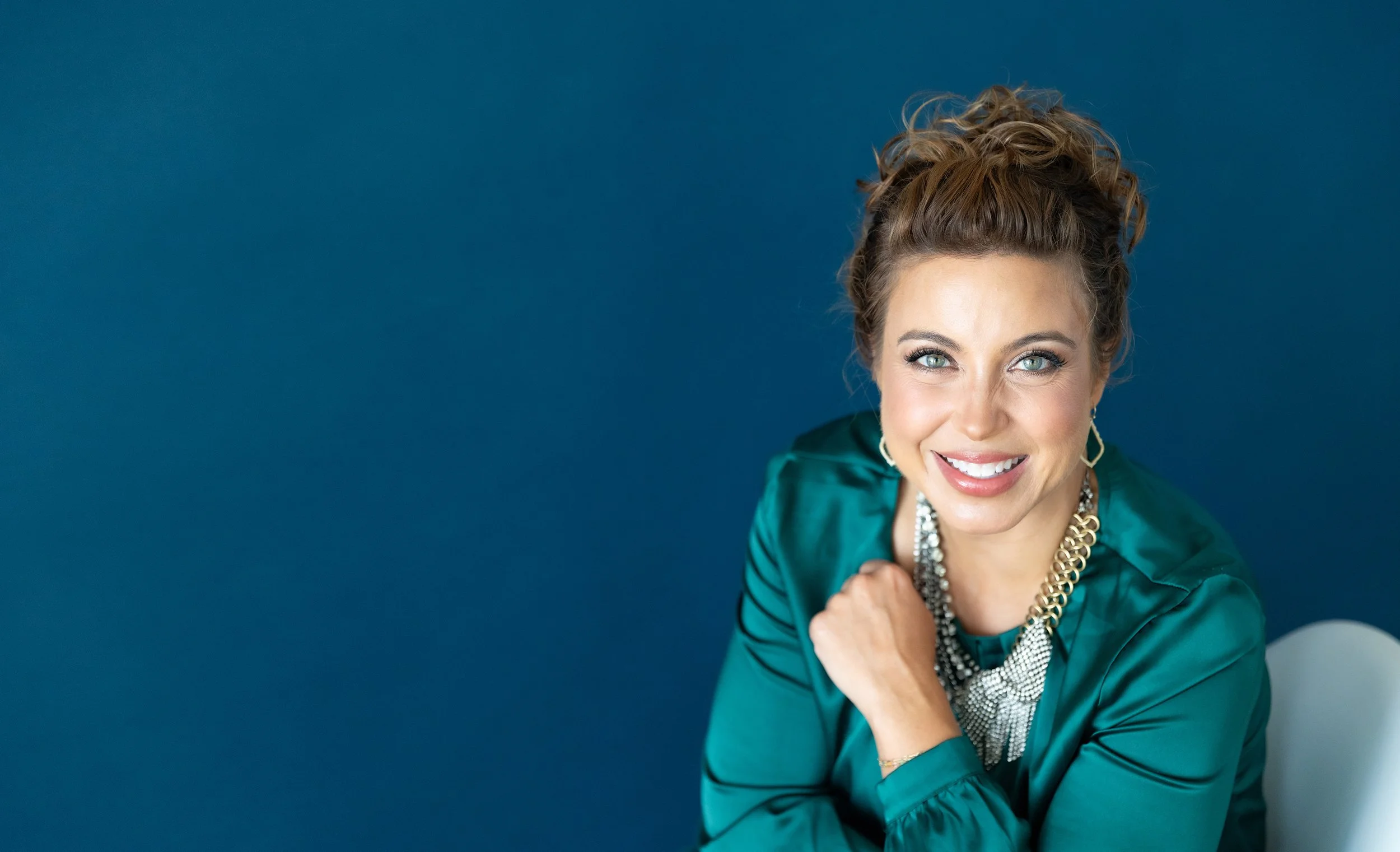 Smiling woman with curly hair wearing teal satin blouse, statement jewelry, and hoop earrings, posing against a blue background.