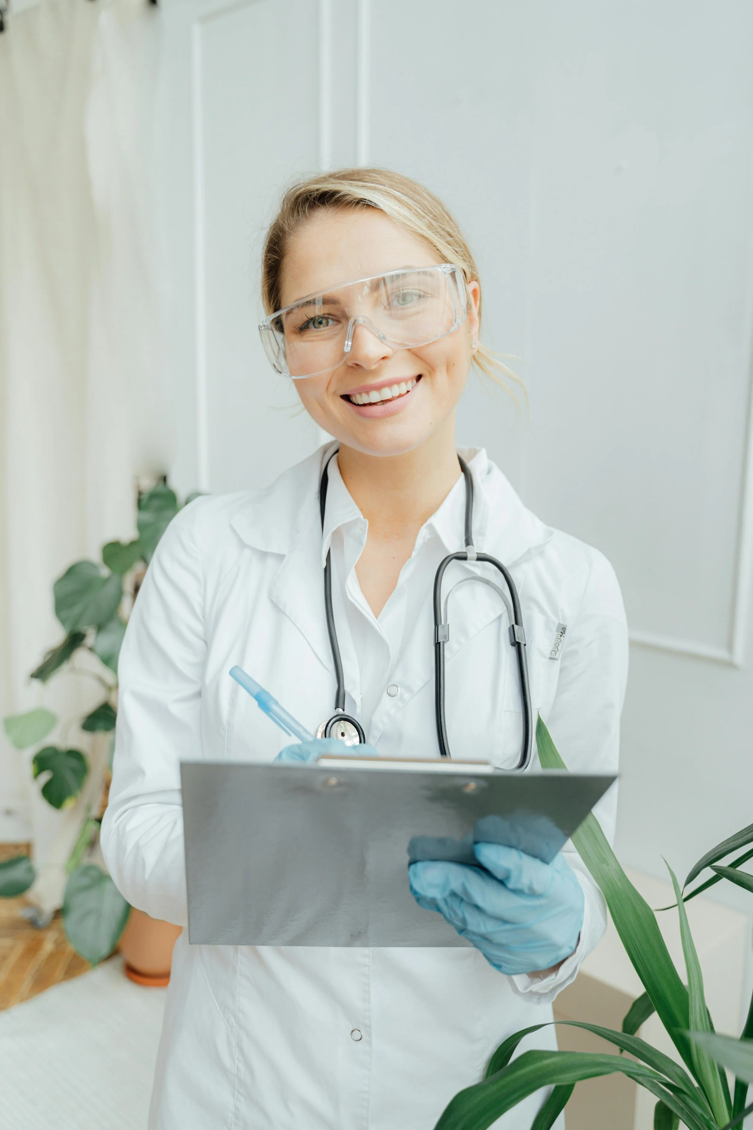 A smiling female doctor with glasses, wearing a white coat and stethoscope, holding a clipboard in a healthcare setting.