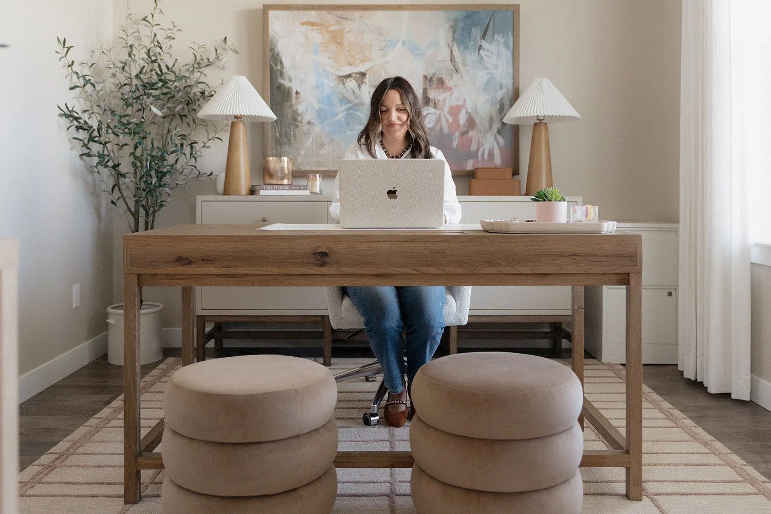 A woman sitting at a desk in a well-decorated home office, working on a laptop, with a large abstract painting on the wall behind her, two lamps, and potted plants on either side of the desk.