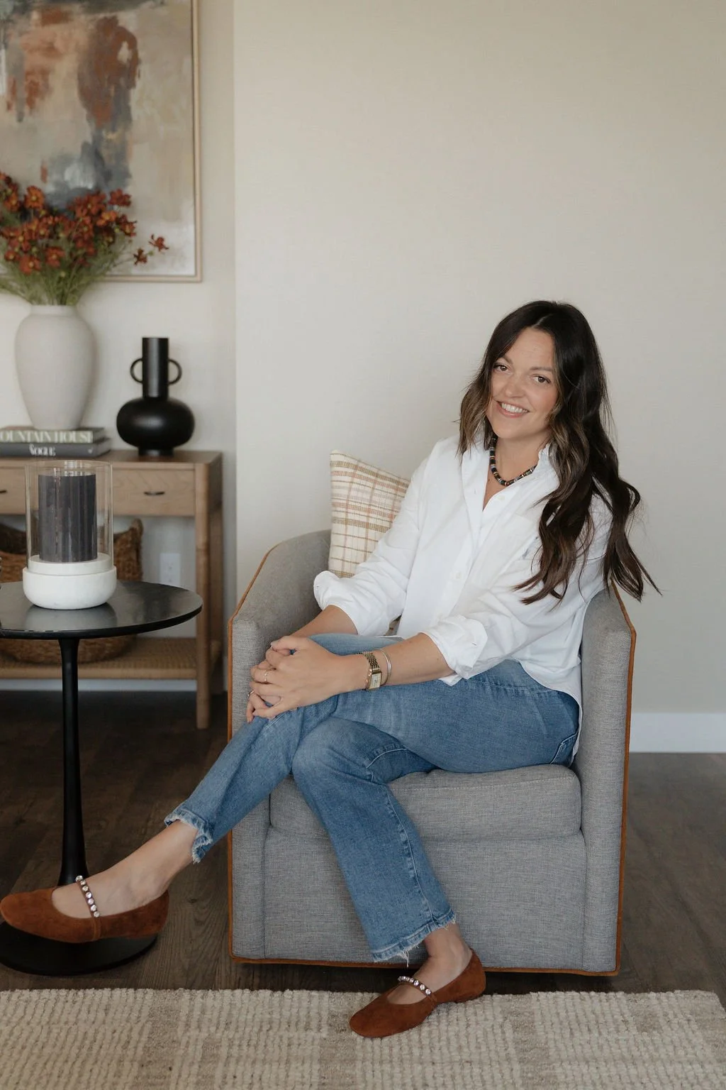 A woman with long dark wavy hair and a white shirt sitting on a gray armchair in a living room, smiling.