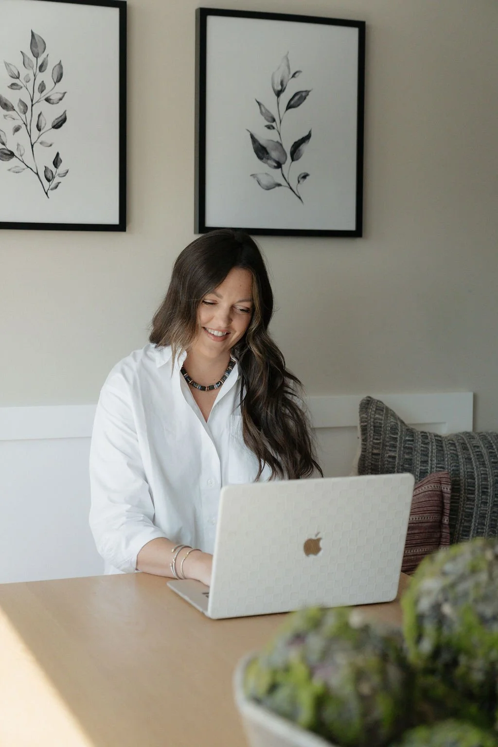 A woman with long dark hair sitting at a wooden table, smiling and looking at a white Apple laptop. Behind her, there are two framed black and white botanical prints on a light-colored wall. There are striped and textured pillows on a bench or bed beside her, and a potted plant in the foreground.