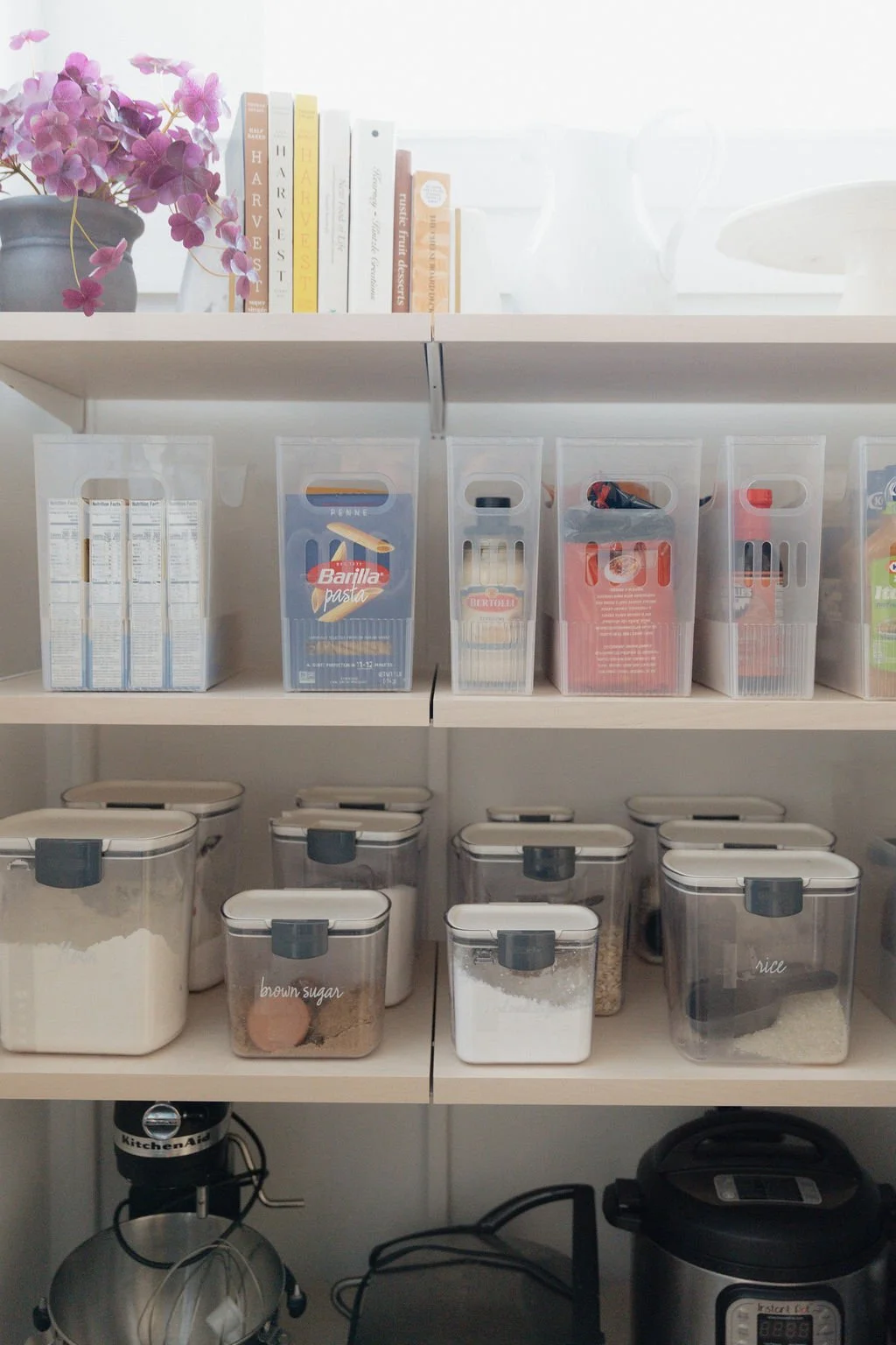 Shelves with books and clear containers of baking ingredients like flour, sugar, and rice, with a stand mixer and a rice cooker below.