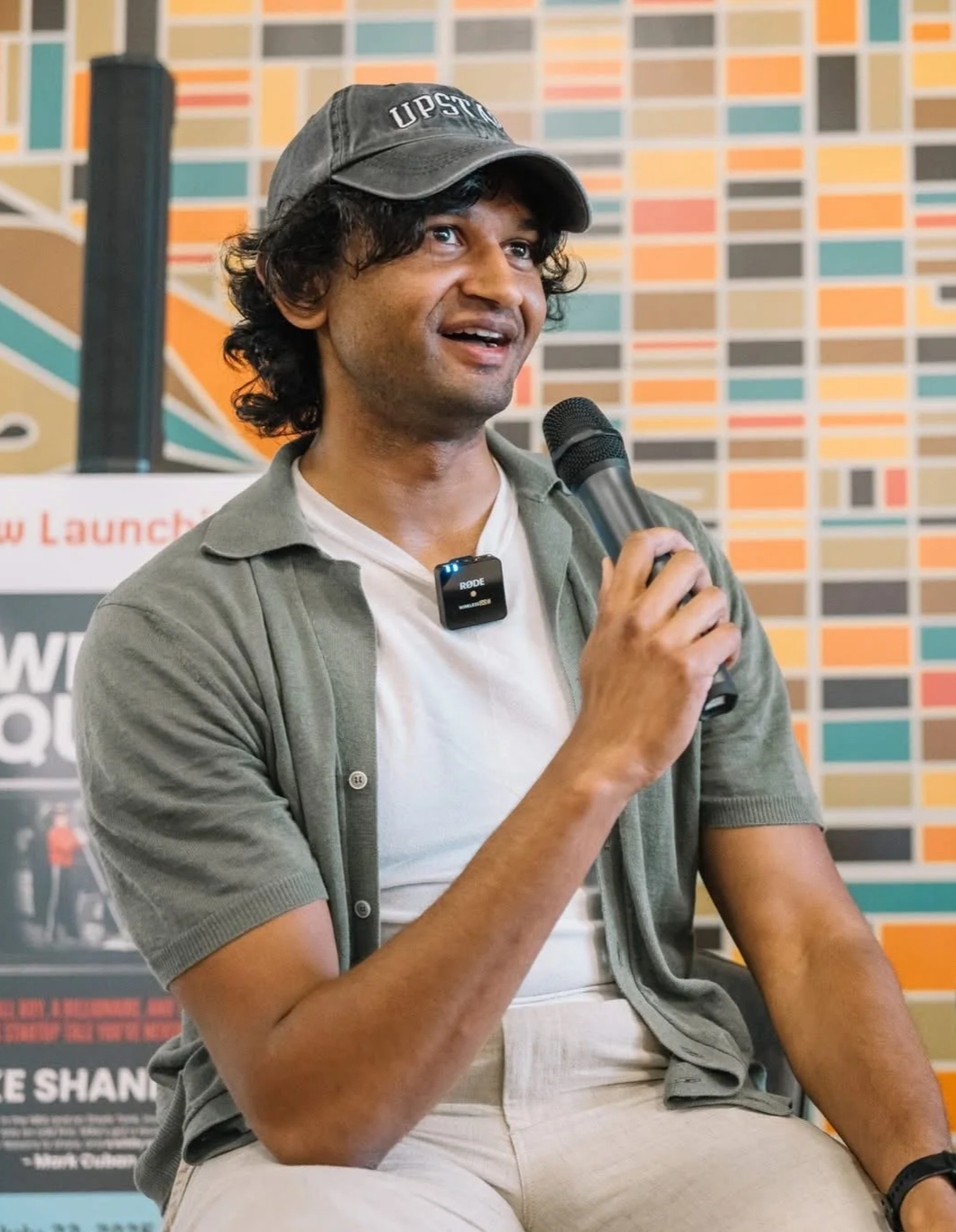 A man speaking into a microphone at an event with a colorful geometric patterned wall in the background.
