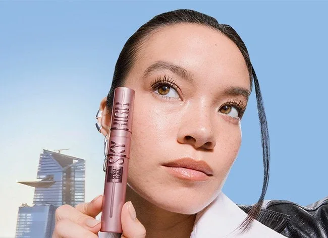 A woman with short blonde hair wearing an orange sleeveless top and beige pants sitting on a bench, writing in a notebook with a black pen, in a room with a potted plant and abstract artwork, sunlight casting shadows on the wall.