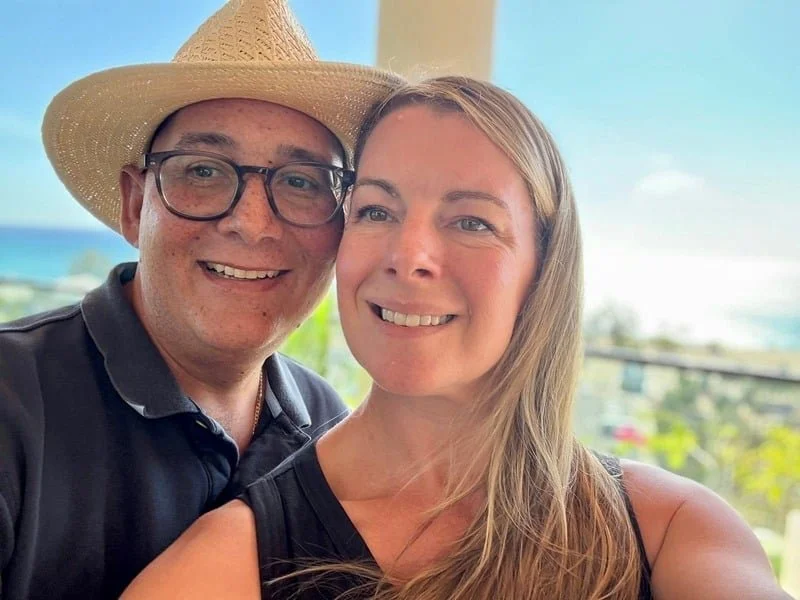 A smiling couple taking a selfie outdoors, with the ocean and a partly cloudy sky in the background, the man wearing a straw hat and glasses, the woman with long blonde hair.