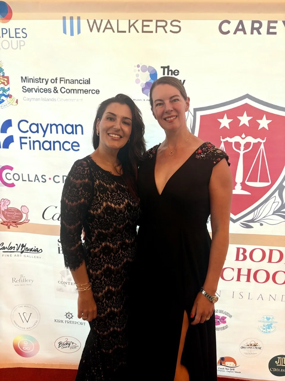 Two women in elegant black dresses smiling at an event, with a backdrop featuring logos and text.