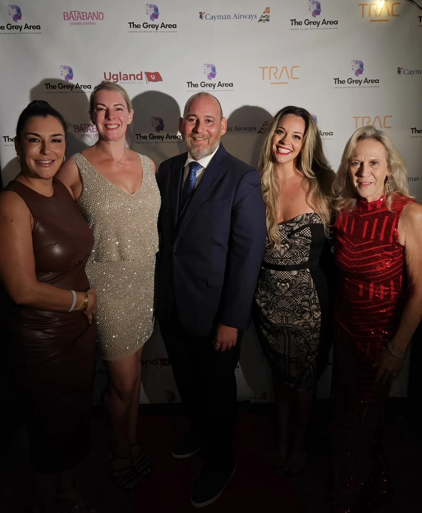 Group of five people, four women and one man, dressed in formal and evening attire, standing in front of a step and repeat backdrop with logos of 'The Grey Area,' 'BalaBano Cayman Carnival,' 'Cayman Airways,' 'Upland Properties,' and 'TRAC', smiling 