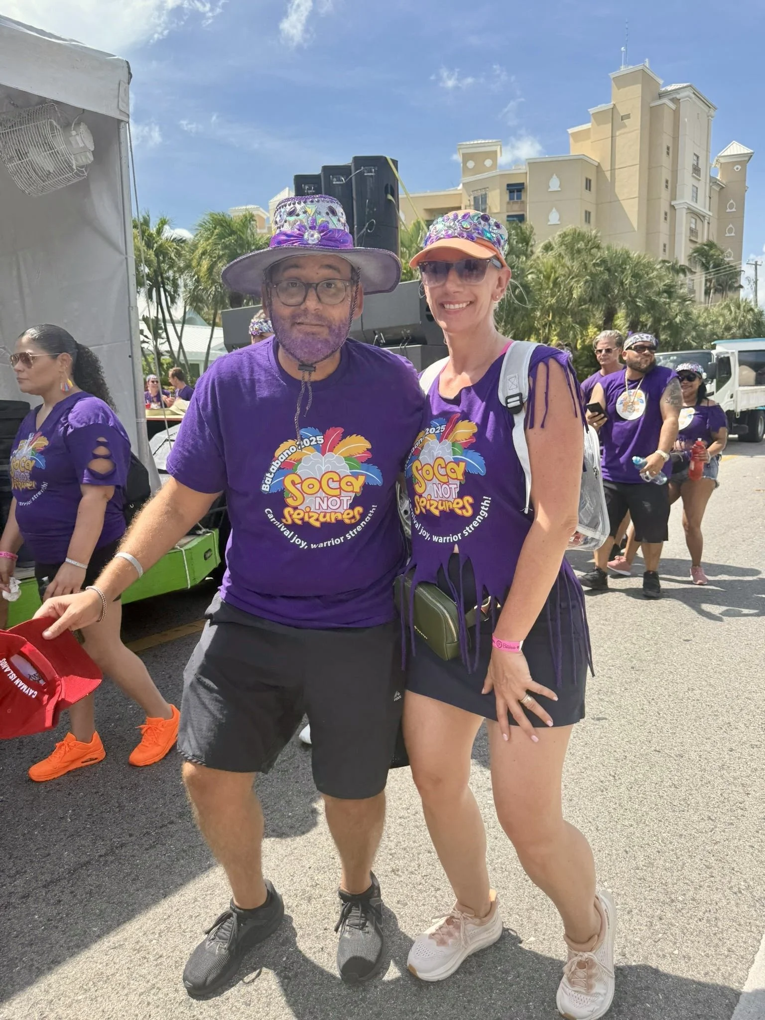 Two people, a man and a woman, standing outdoors at a festival or event, both wearing purple T-shirts with colorful text and designs, smiling at the camera. The man wears a purple hat decorated with shiny embellishments, black shorts, and glasses; th