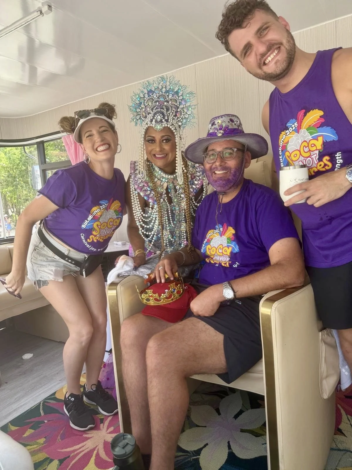 Four people wearing purple shirts with a colorful event logo, one person in elaborate costume and headdress, and two men with glasses and hats, smiling indoors with a window and greenery outside.