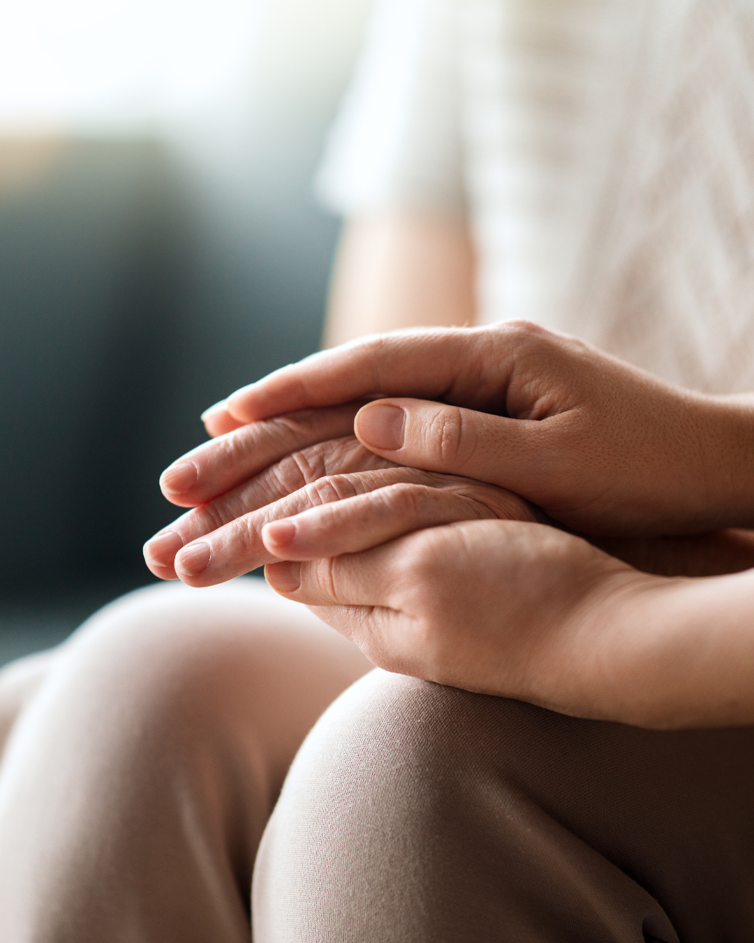 Close-up of a person's hands resting on their lap, with fingers gently intertwined, wearing beige clothing.