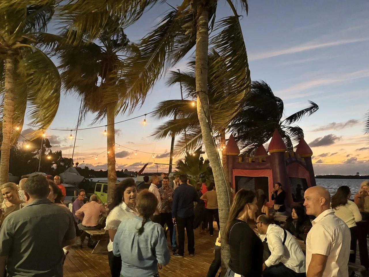 People gathered outdoors at sunset near palm trees, with a small inflatable castle, string lights, and a boat in the background.