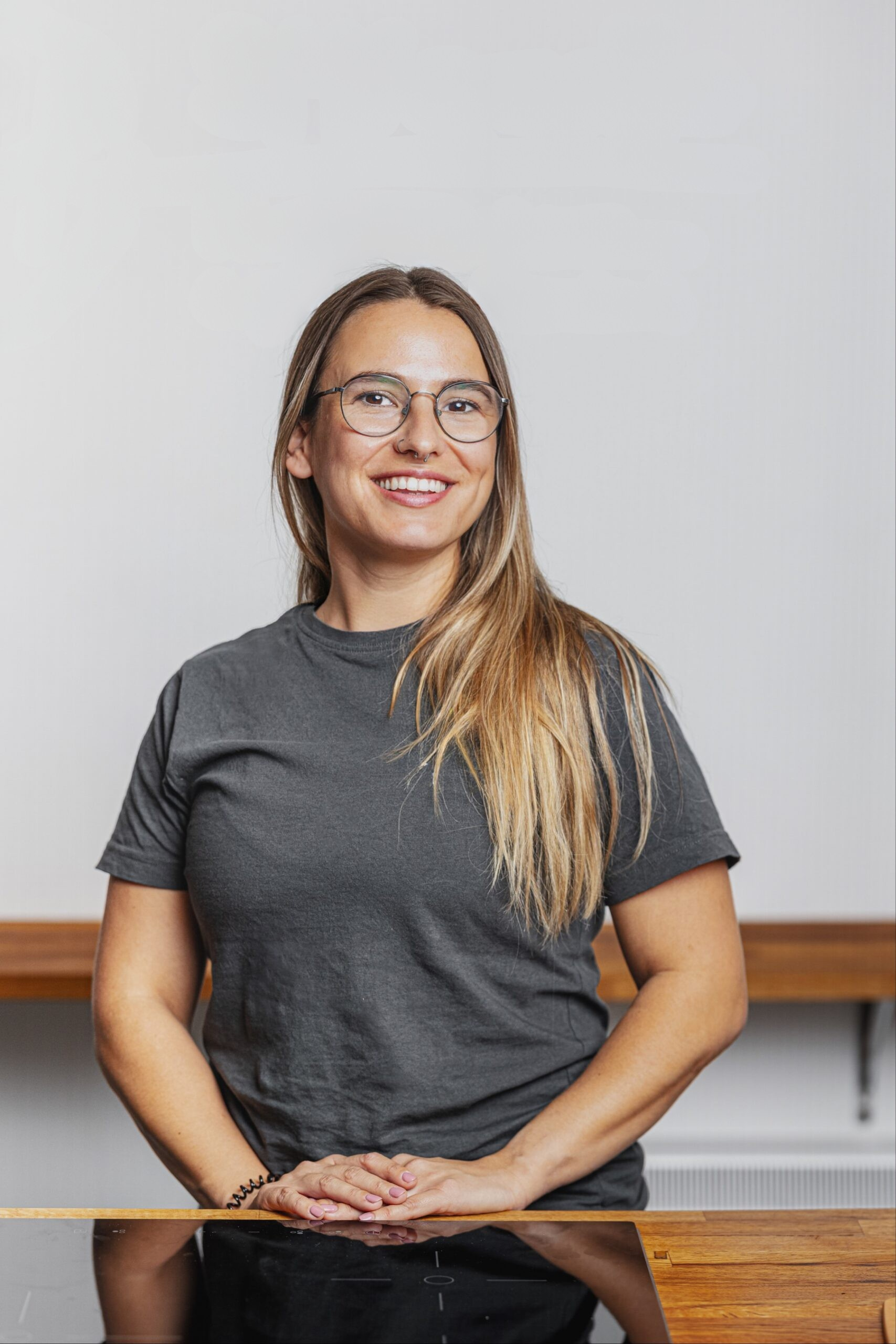 A woman with long blonde hair, glasses, and a gray t-shirt, smiling while standing in front of a white wall and wooden table.
