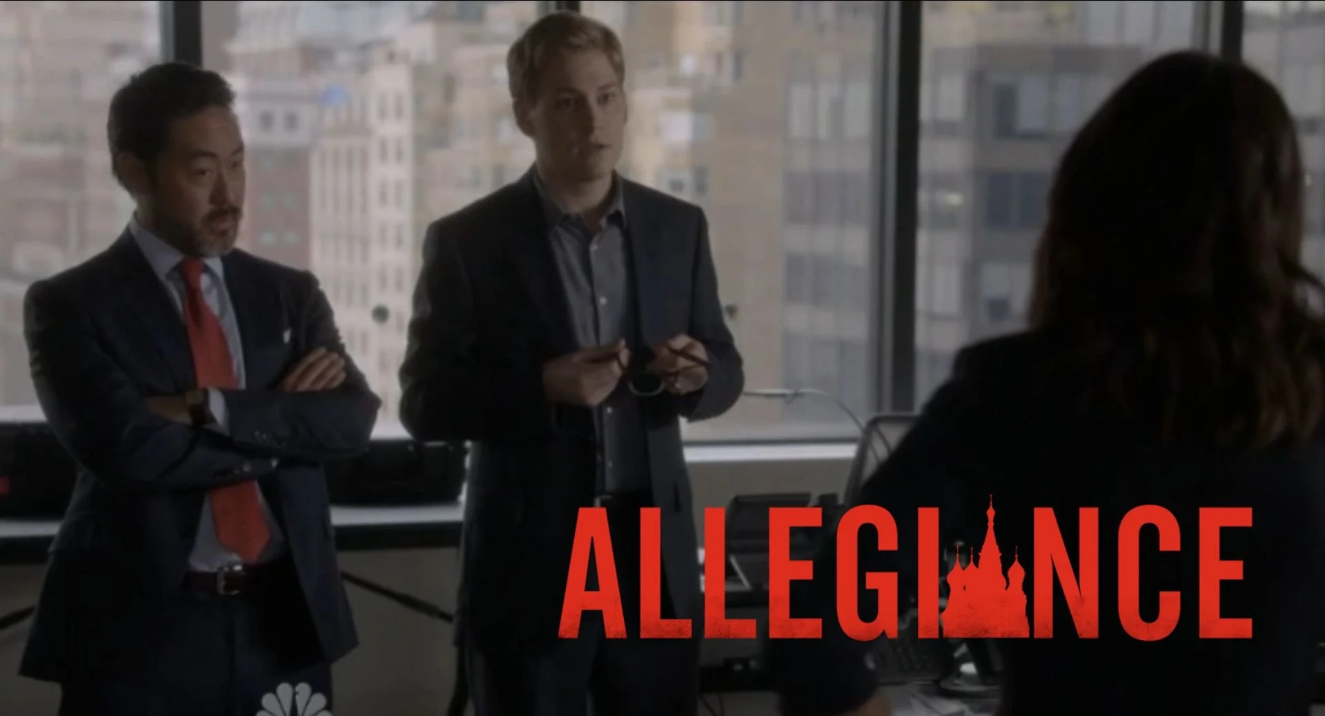 Two men standing in an office, engaged in conversation, with a woman sitting at a desk in front of them, and a cityscape outside the window. The word "ALLEGANCE" with a silhouette of a castle is displayed at the bottom.