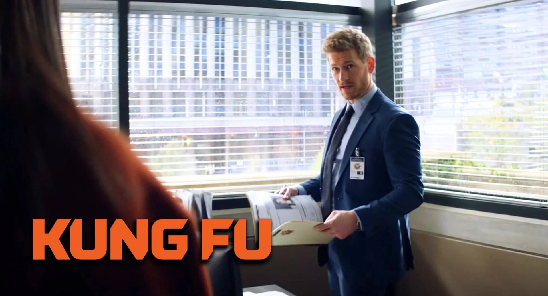 A man in a blue suit and tie with a security badge on his chest is standing next to a window, holding open a magazine or brochure, inside an office or security booth with horizontal blinds. There is another person partially visible in the foreground,