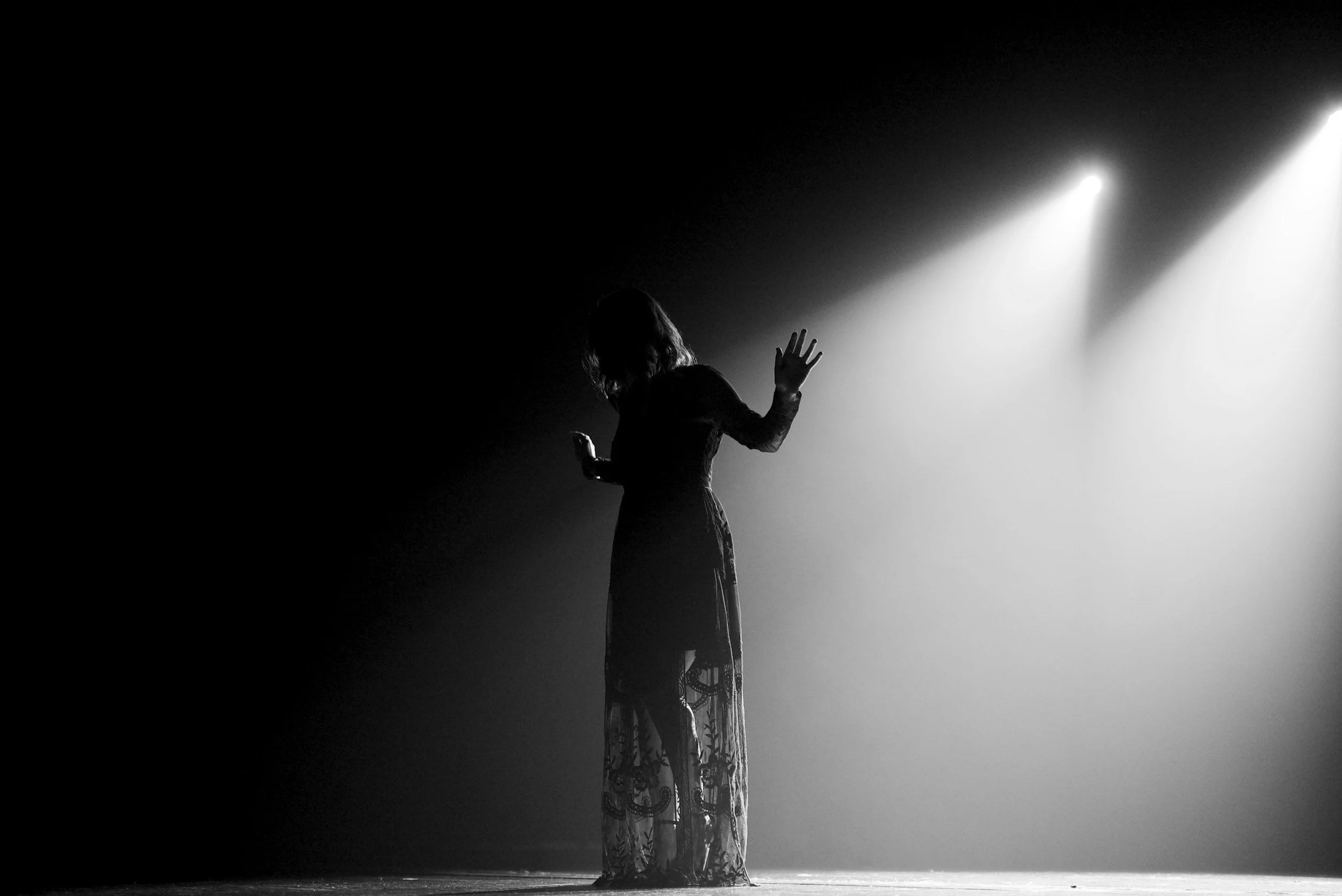 A silhouette of a woman standing on stage with her back to the camera, illuminated by bright stage lights, wearing a long lace dress.