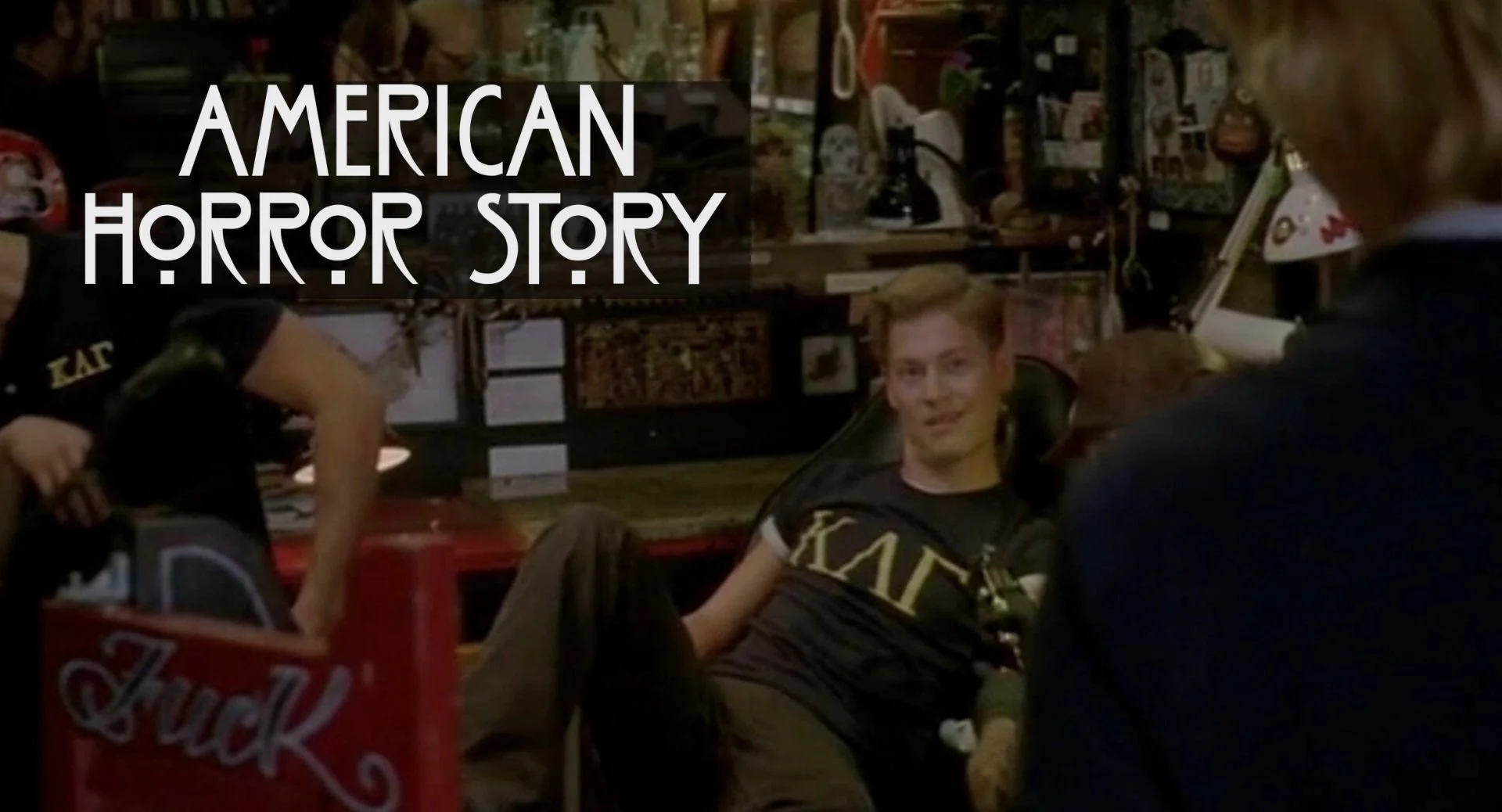 A young man sitting on a chair, in a store decorated with various collectibles and antiques, with a person in the foreground and a woman in the background.