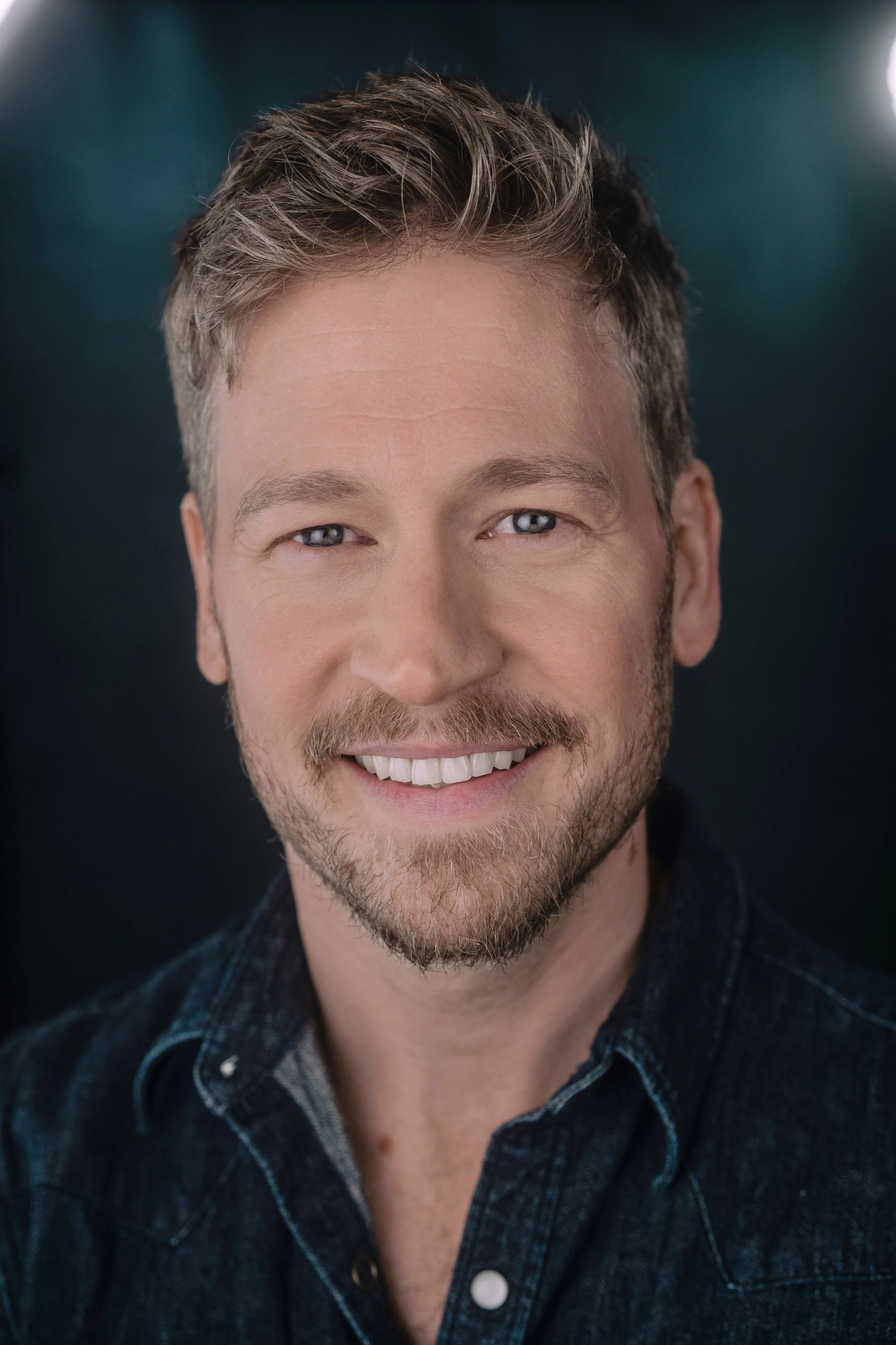 A smiling man with light skin, blonde hair, and a beard, wearing a dark blue button-up shirt, against a dark background.