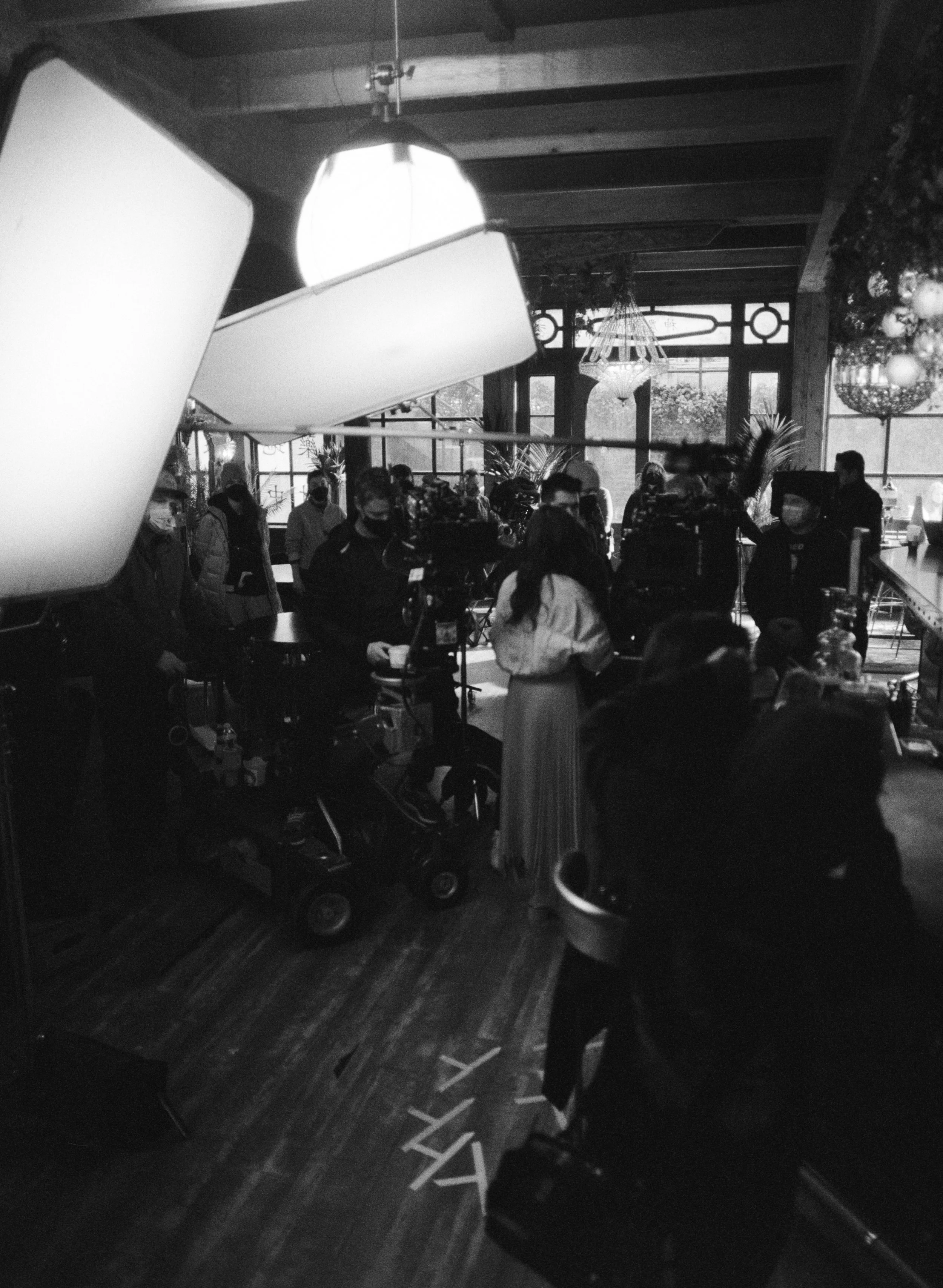 Film crew filming inside a bustling restaurant or café with large windows, chandeliers, and hanging lights, with people gathered around and working.