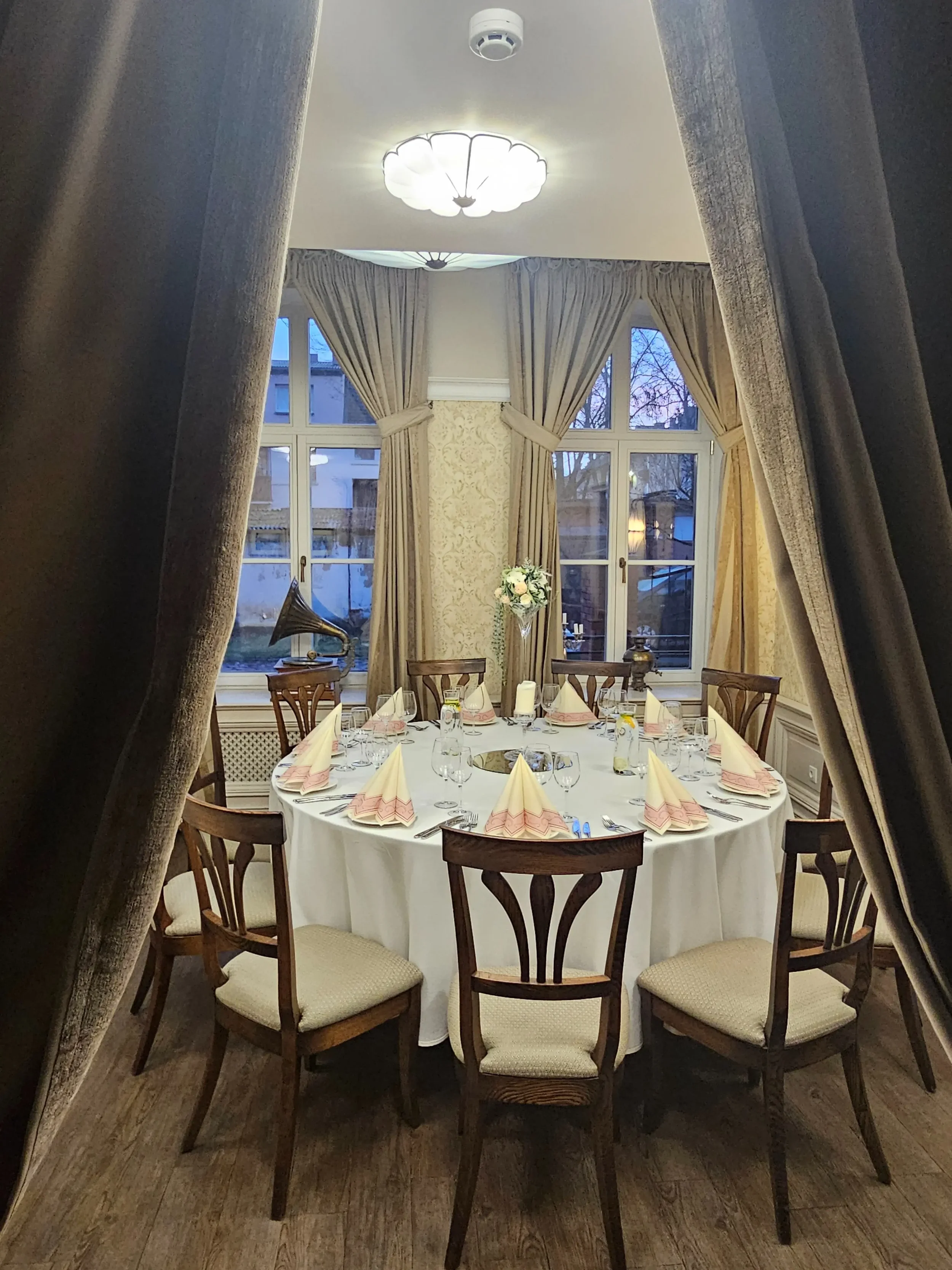 Elegantiška restorano salė su apvaliu stalu, žvakėmis ir antikvariniu gramofonu fone.