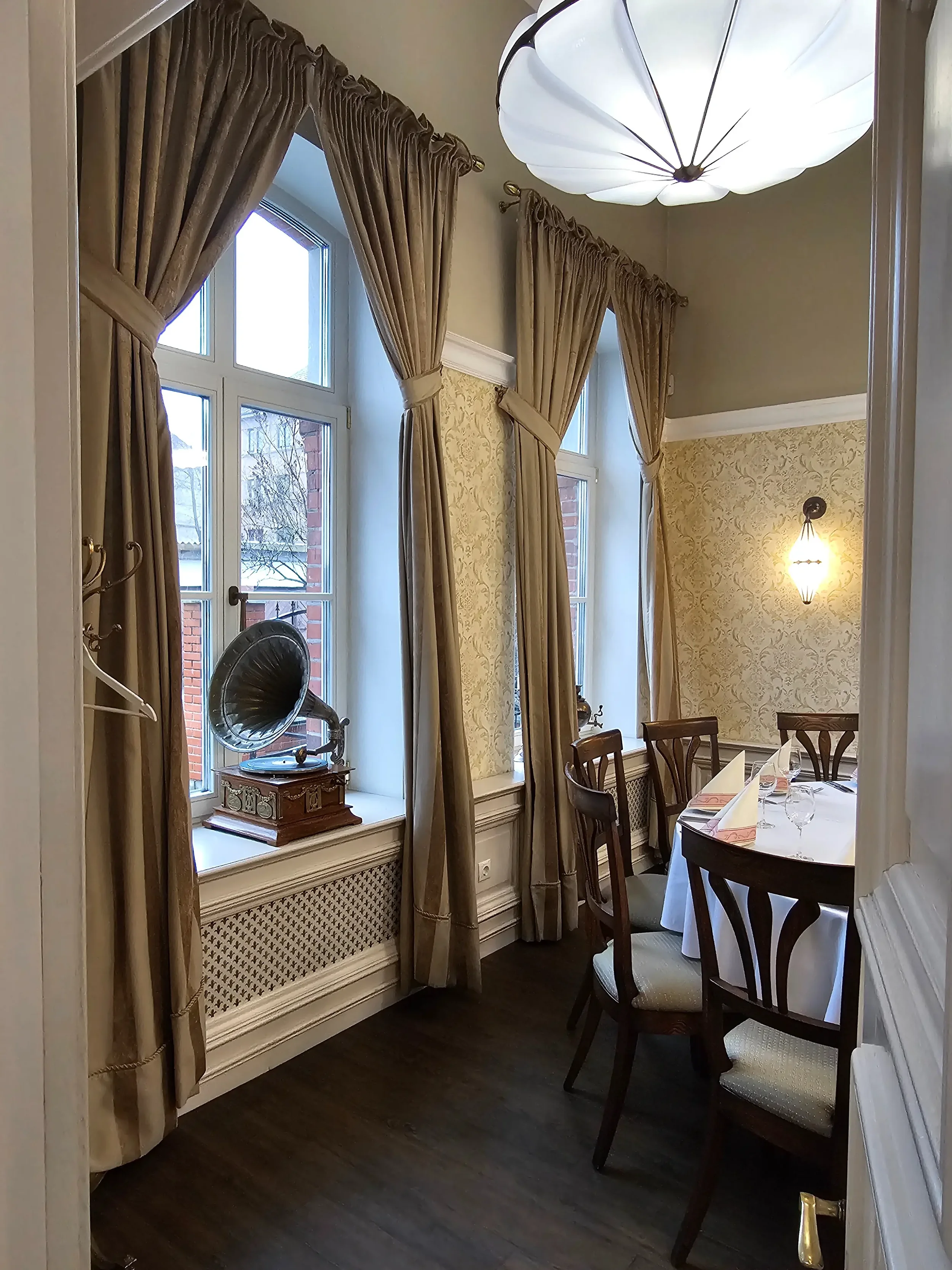 Klasikinio stiliaus restorano erdvė su apvaliu stalu, dideliais langais ir antikvarinėmis detalėmis.