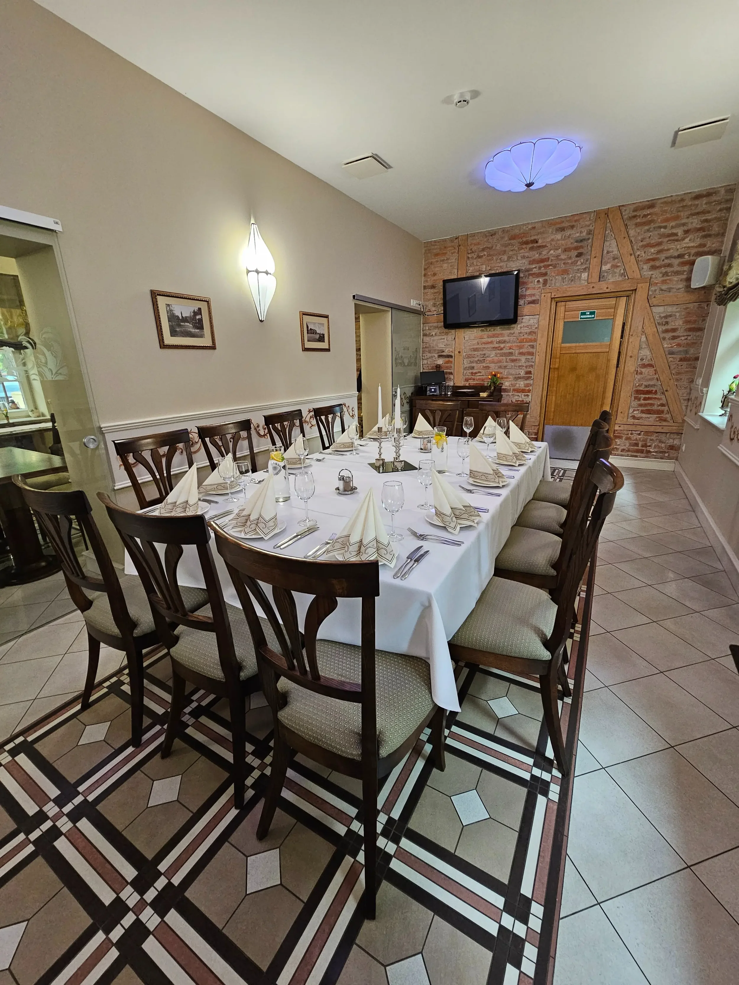 Ilgas banketinis stalas, paruoštas šventei privačioje restorano erdvėje su plytų siena.