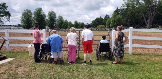 Six people, including two in wheelchairs, stand by a white fence watching a horse arena on a cloudy day.
