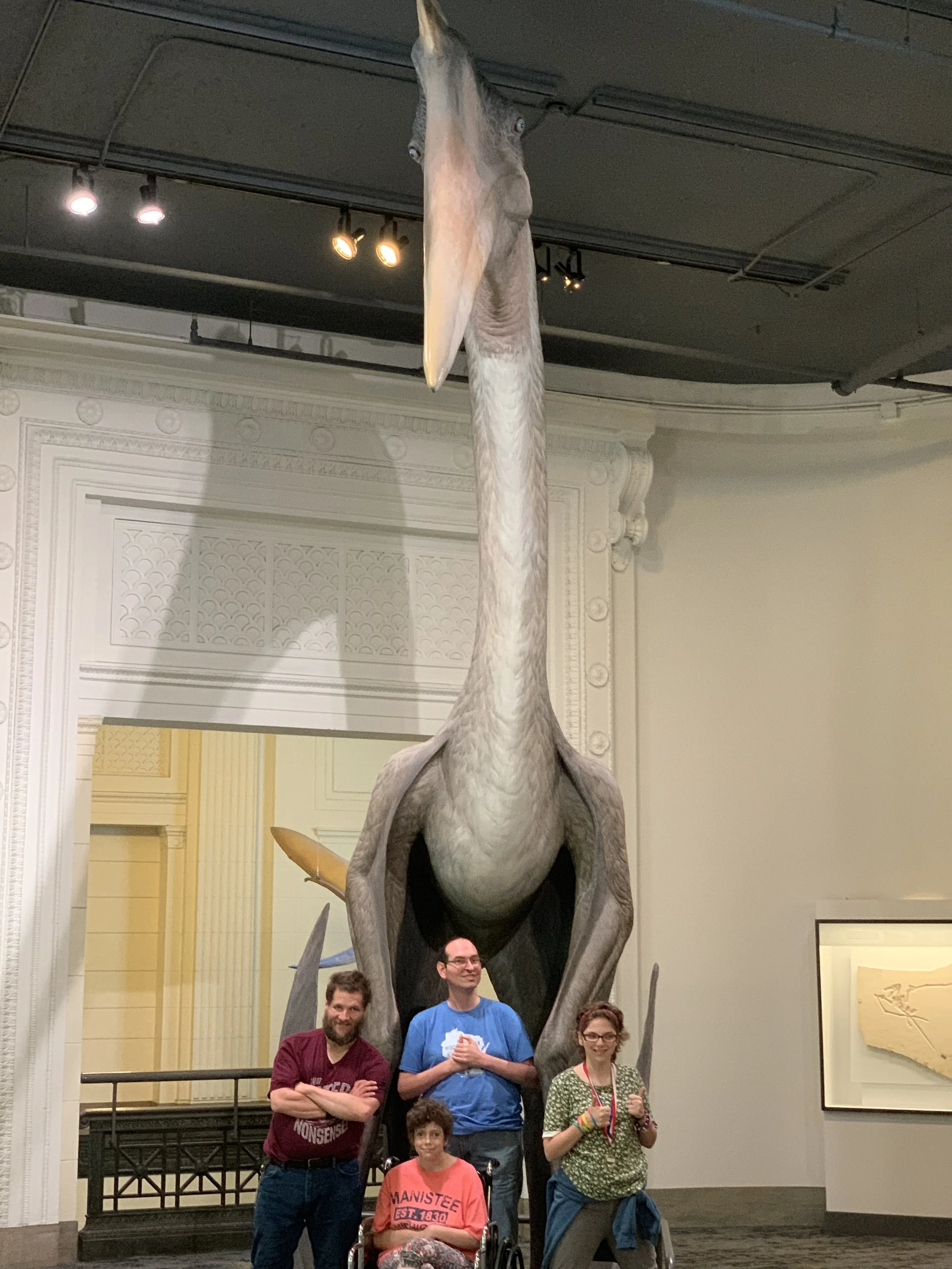 Four people, including a young girl in a wheelchair, posing in front of a large blue whale skull exhibit in a museum. The group is smiling with the whale's jaw open behind them.